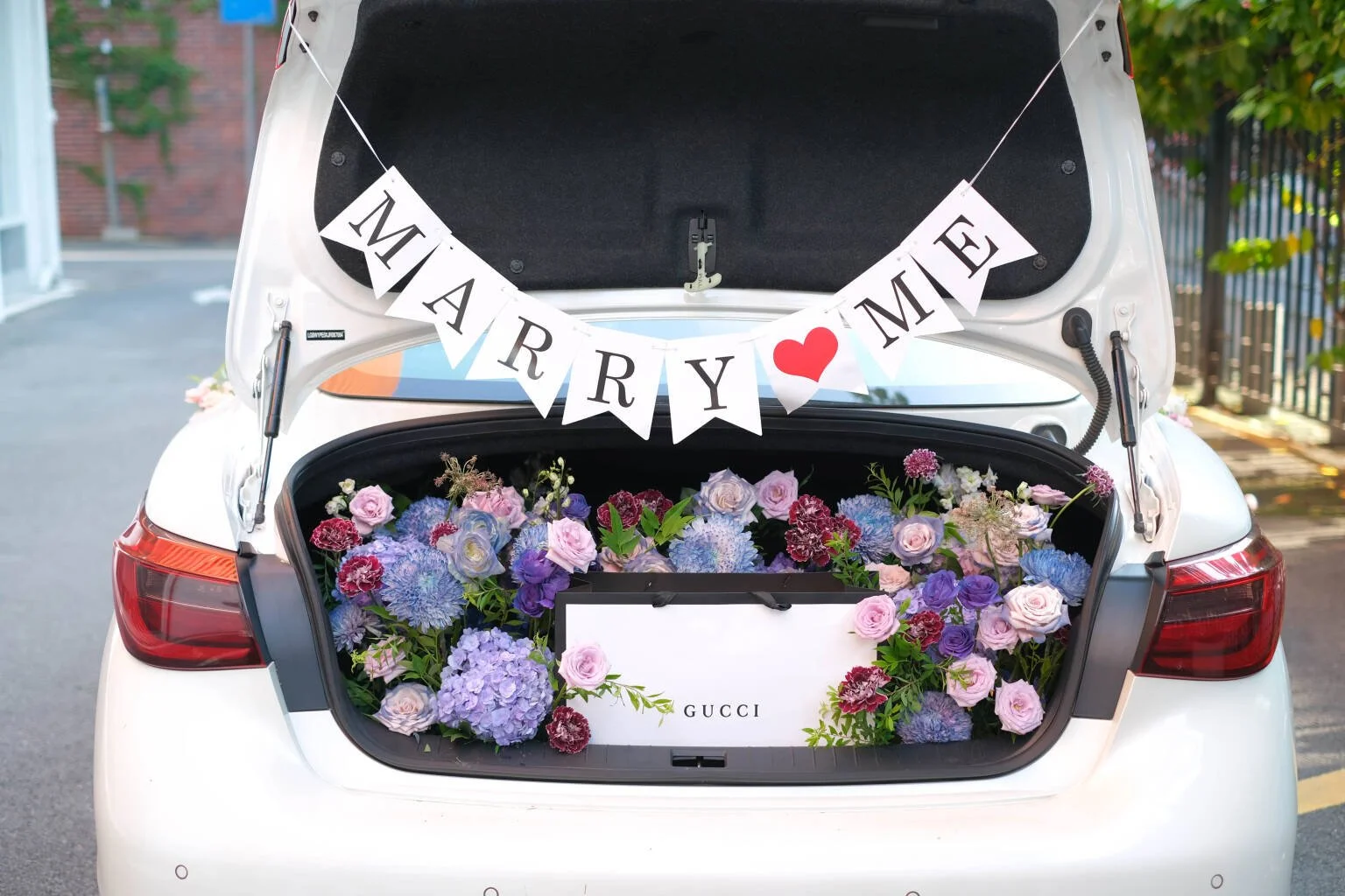 The open trunk of a white car filled with pink, purple, and blue flowers, decorated with a 'Marry Me' banner and a white box labeled 'GUCCI'.