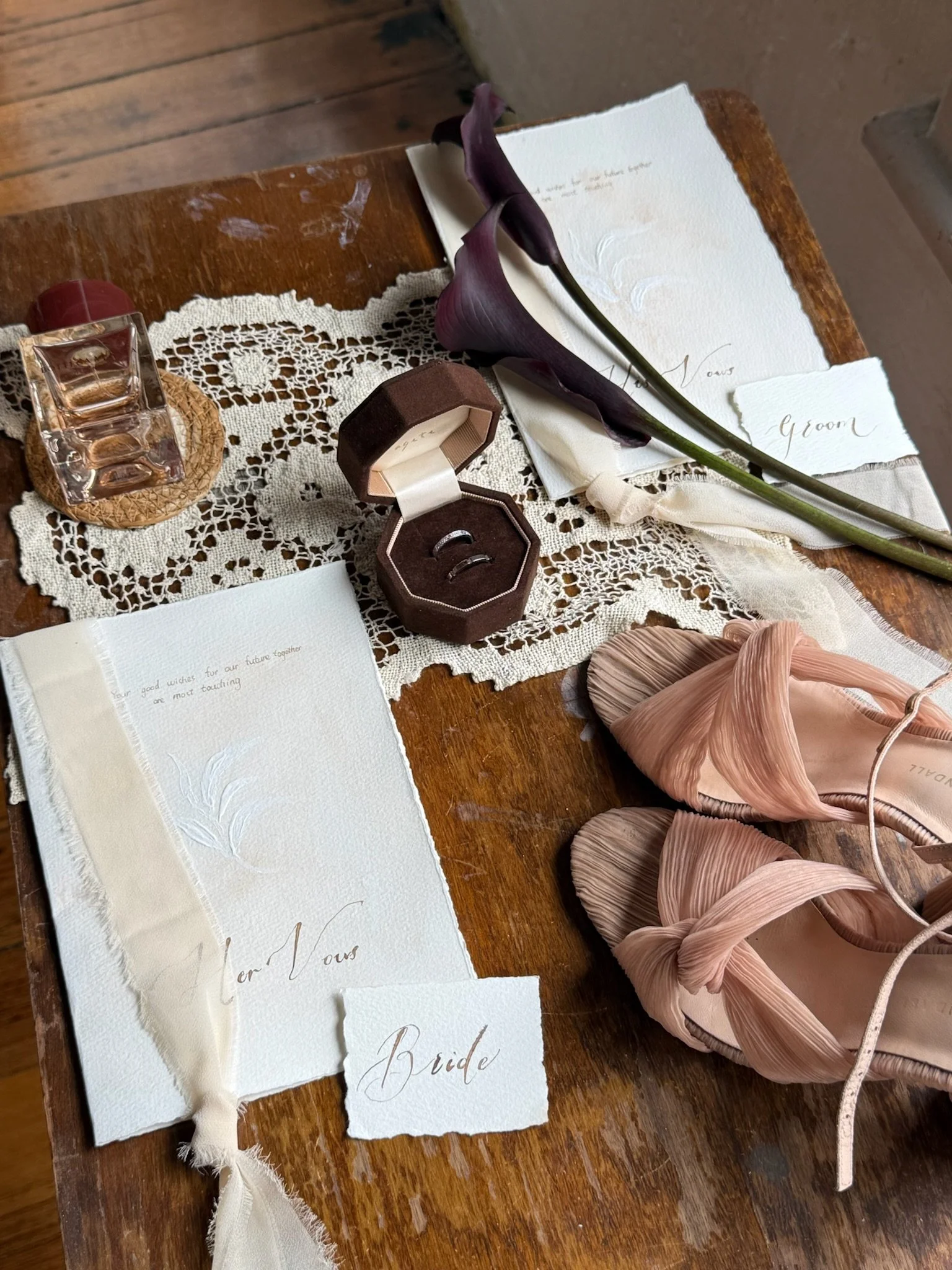Wedding items on a wooden table including a lace doily, a bottle of perfume, a ring box with rings, dark purple calla lilies, blush pink high heel shoes, and handwritten cards reading 'Bride' and 'Dear Vous'.