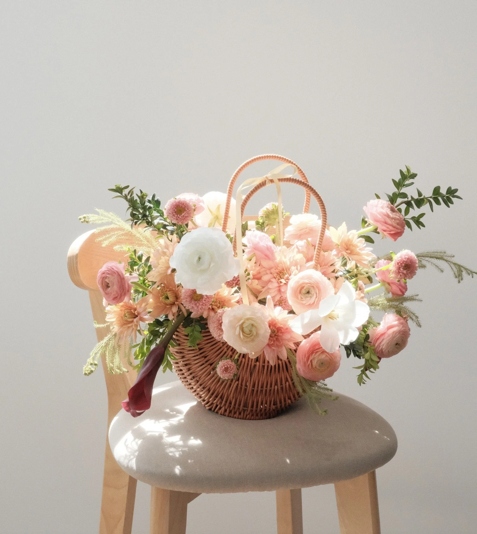 A wicker basket filled with pink and white flowers, sitting on a beige cushioned stool against a plain off-white wall.