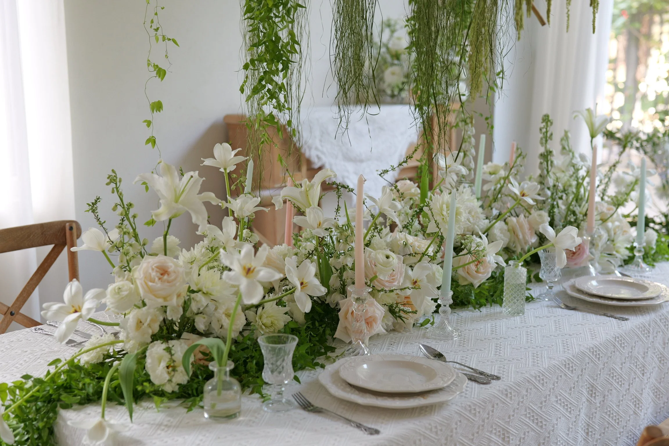 Elegant dining table decorated with white and blush flowers, candles, and tableware, set for a special occasion in a bright room with greenery.