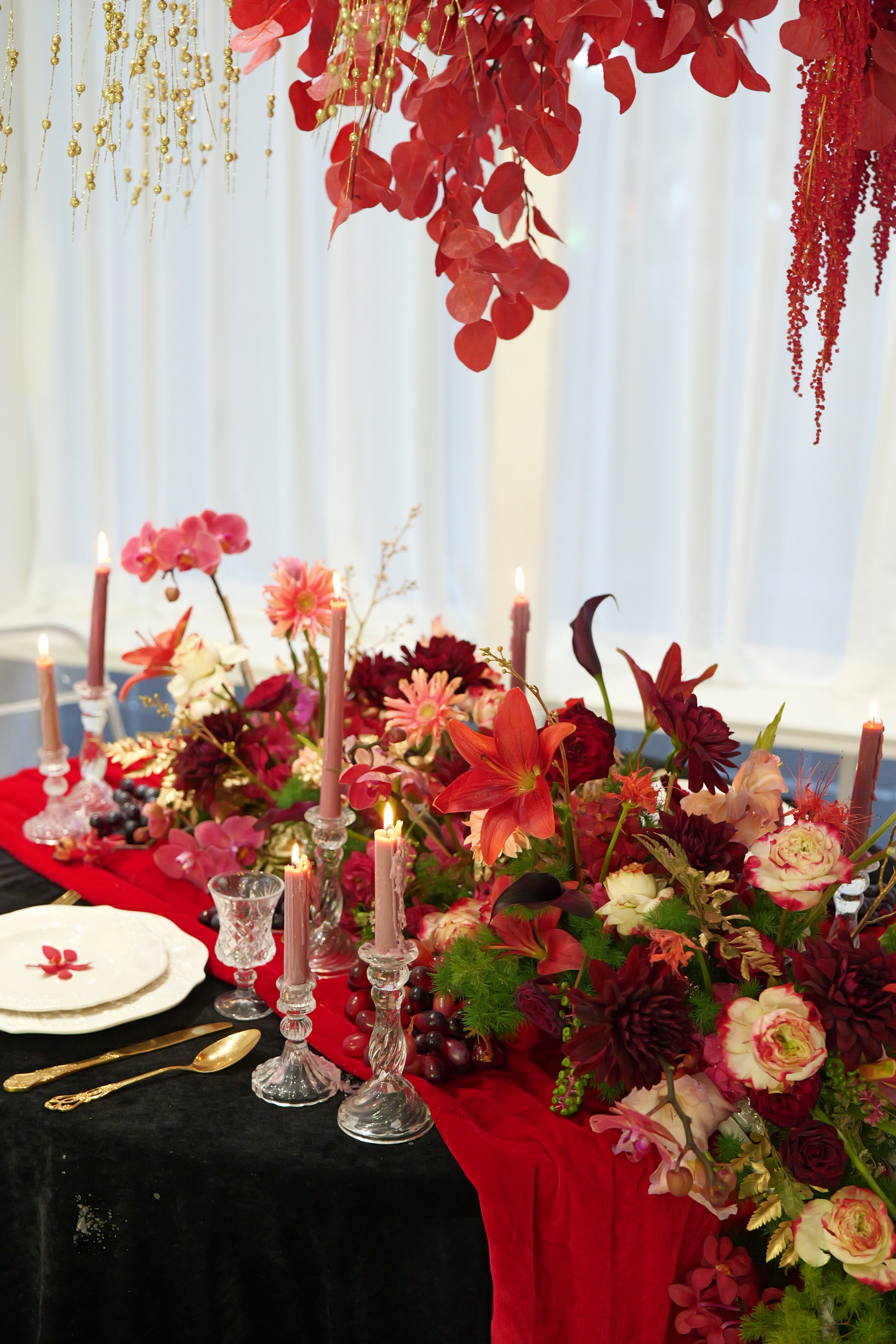 A decorated dining table with a black tablecloth, a red runner, and a large floral centerpiece with pink, red, and white flowers, surrounded by pink candles, gold cutlery, and delicate glassware, with hanging red and gold decorations overhead.