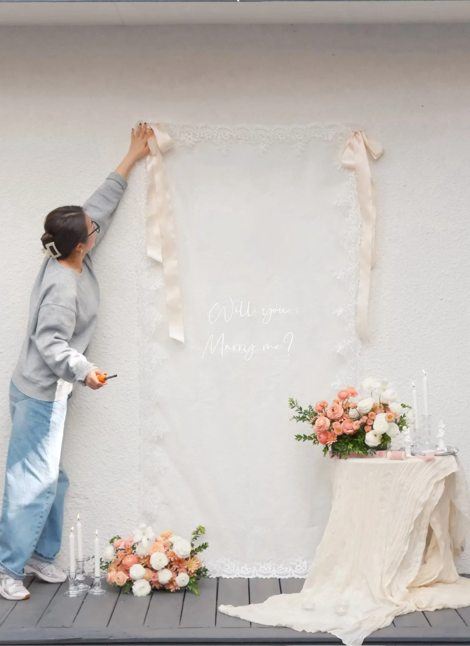 A woman is hanging a lace and satin ribbon banner above a white backdrop with the words 'Will you Marry me?' written on it, decorated with pink and white flowers and candles on a table.