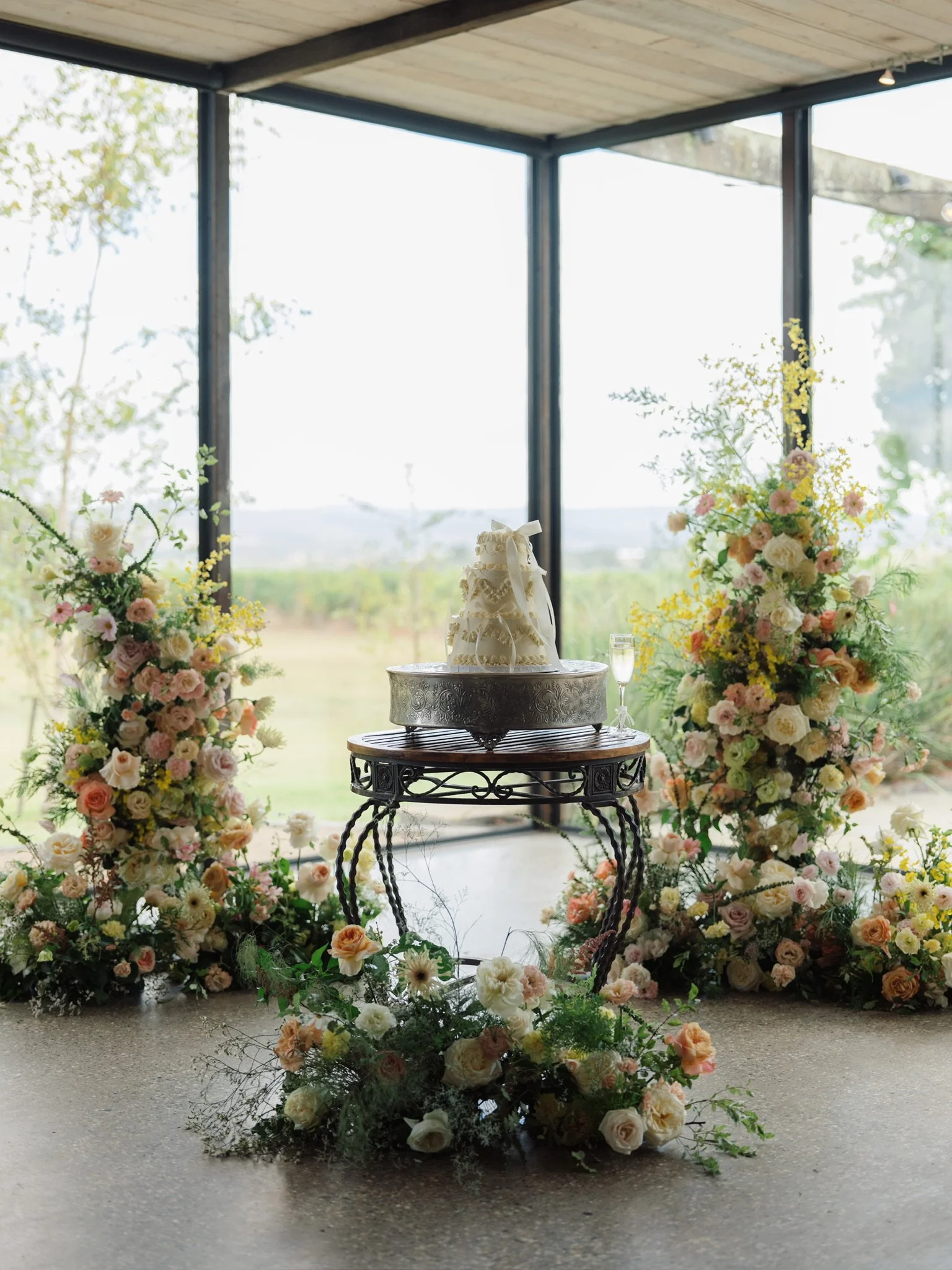 A wedding cake with white icing and lace decoration on an ornate silver stand, surrounded by pastel-colored flowers, in front of large windows revealing a green landscape. A champagne flute is beside the cake.