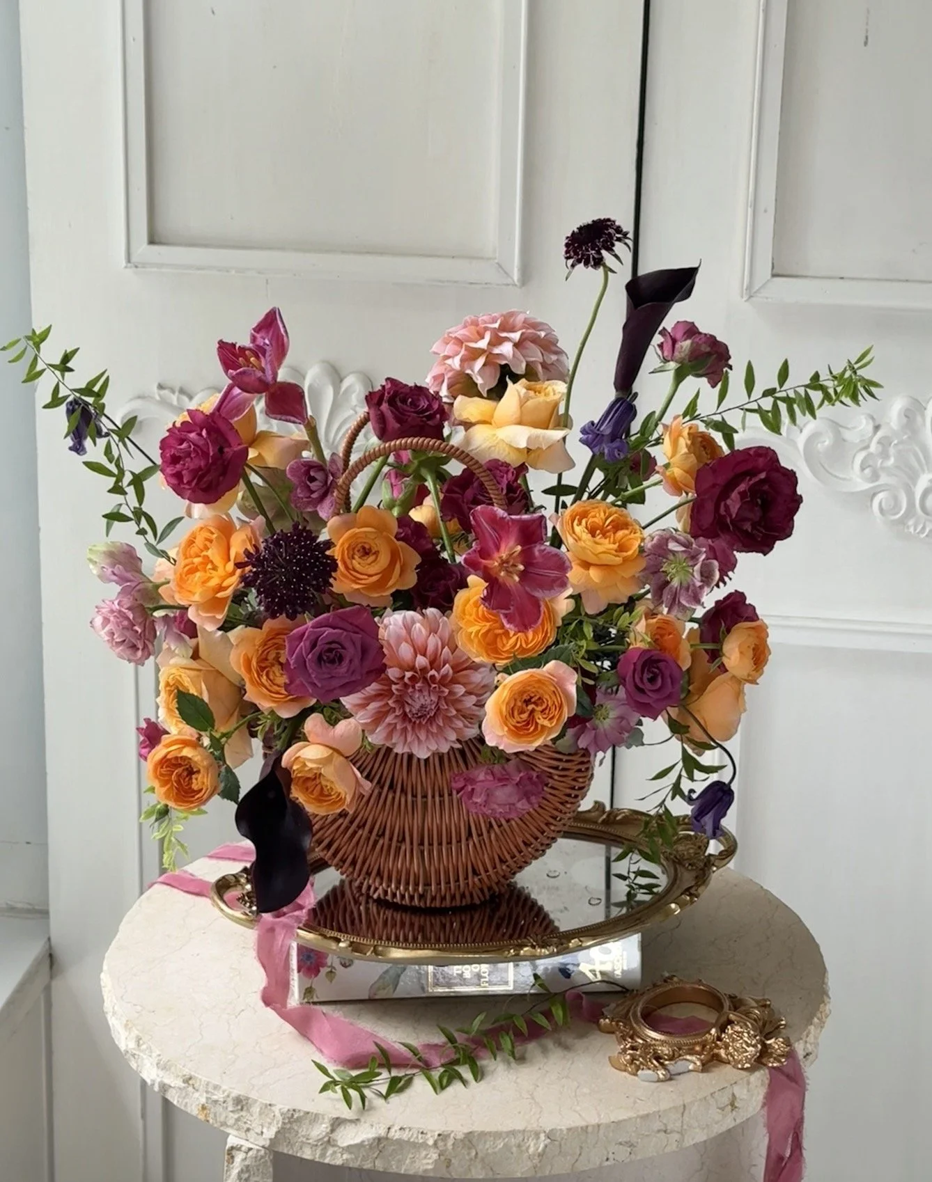 A colorful arrangement of flowers in a woven basket on a marble table, with a decorative mirror and a pink ribbon.