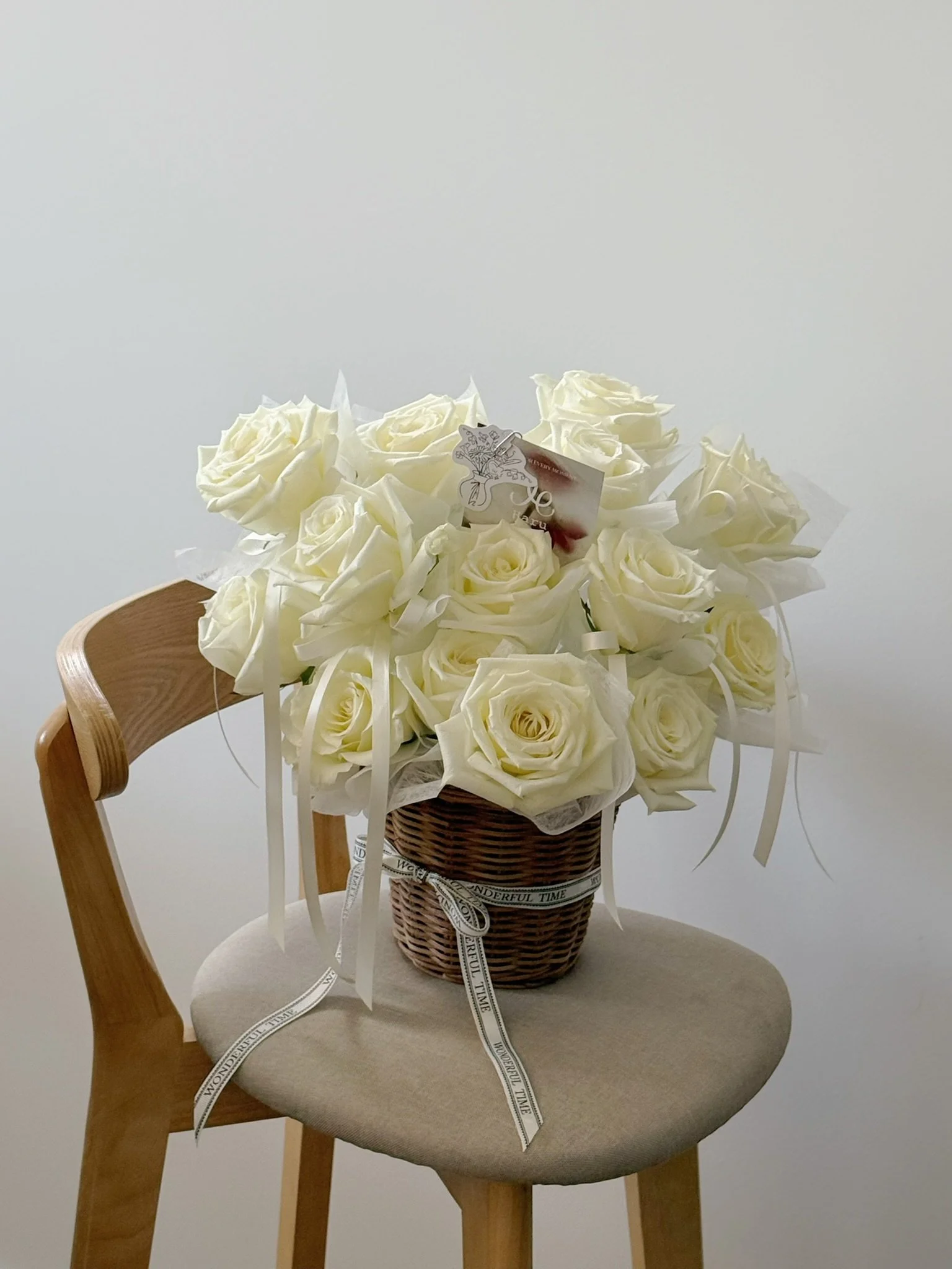 A basket filled with white roses and ribbons, placed on a wooden chair with a beige seat and backrest, against a plain white background.