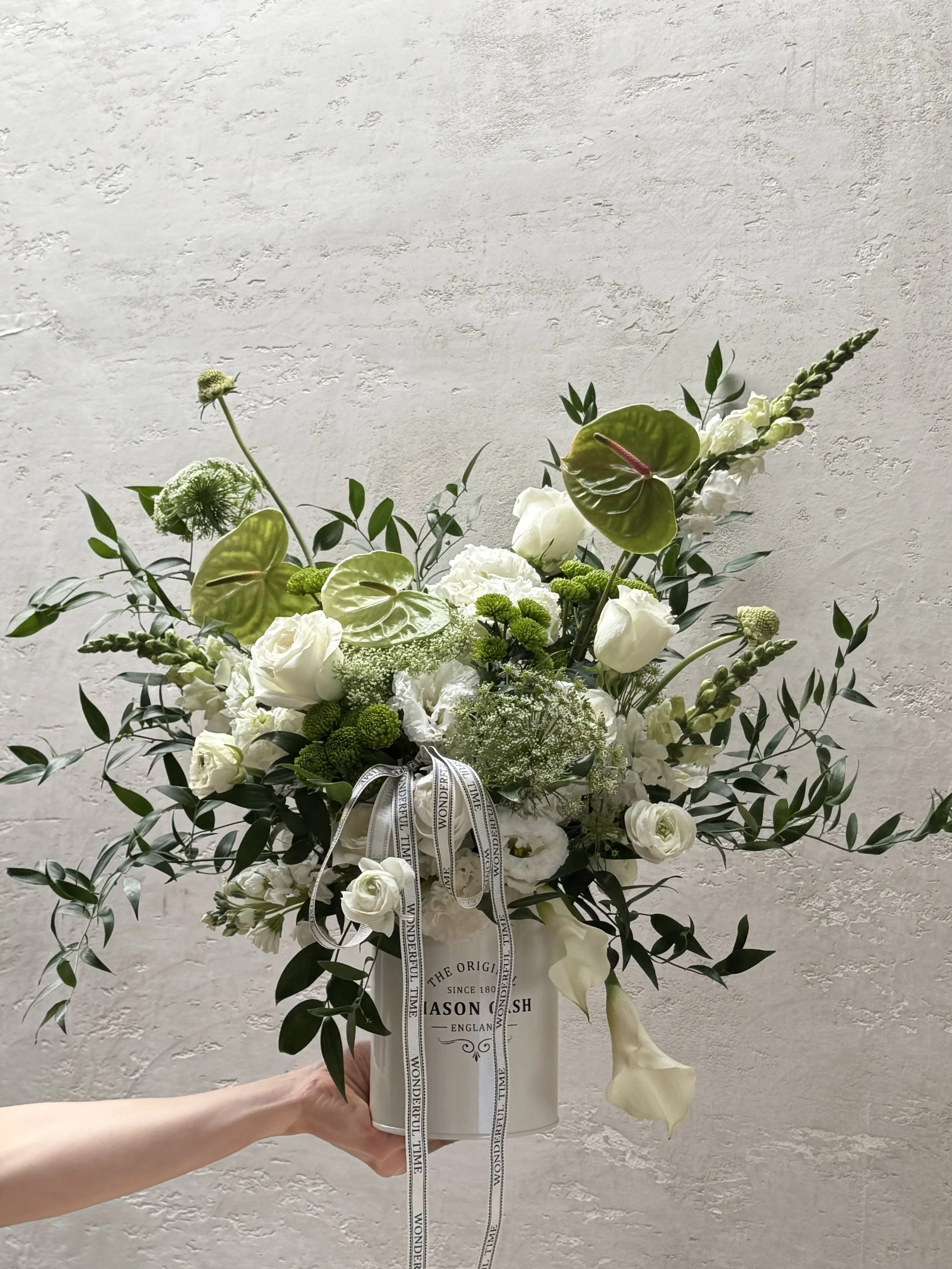 A large white flower arrangement in a white vase with black text, held by a pale hand against a textured white wall. The arrangement contains white roses, calla lilies, anthuriums, and various greenery, decorated with black and white ribbon.