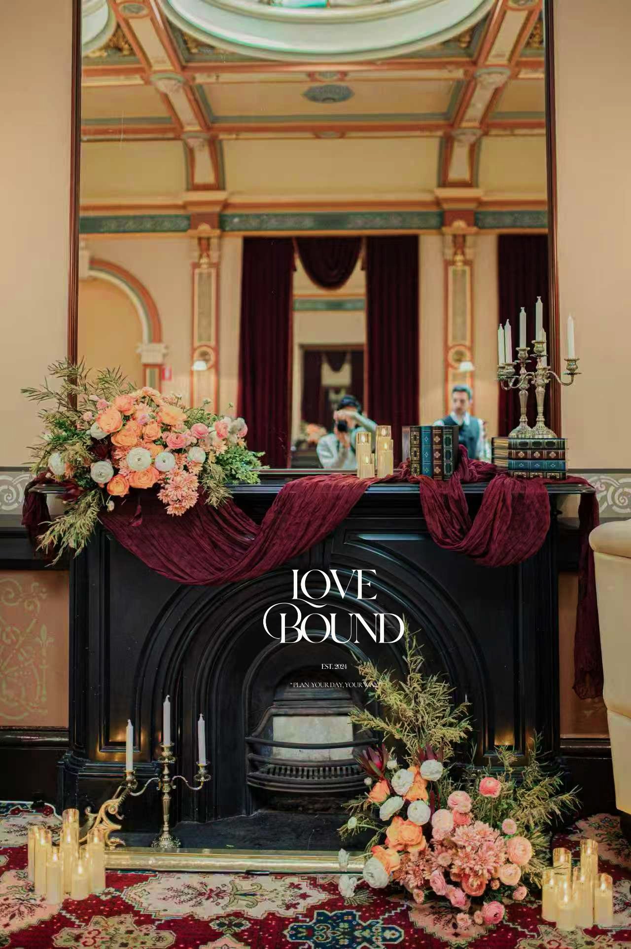 Decorative fireplace with floral arrangements, candles, and books in a vintage-inspired room, with a large mirror reflecting people and detailed ceiling.
