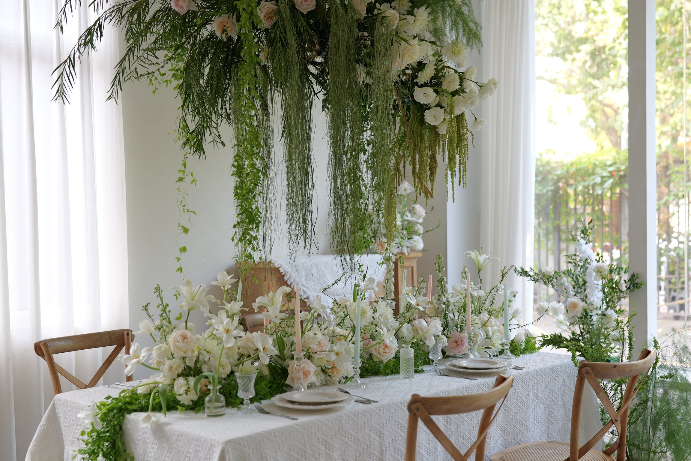 Elegant dining table with white floral arrangements, candles, and greenery centerpiece in a bright room with large windows and sheer curtains.