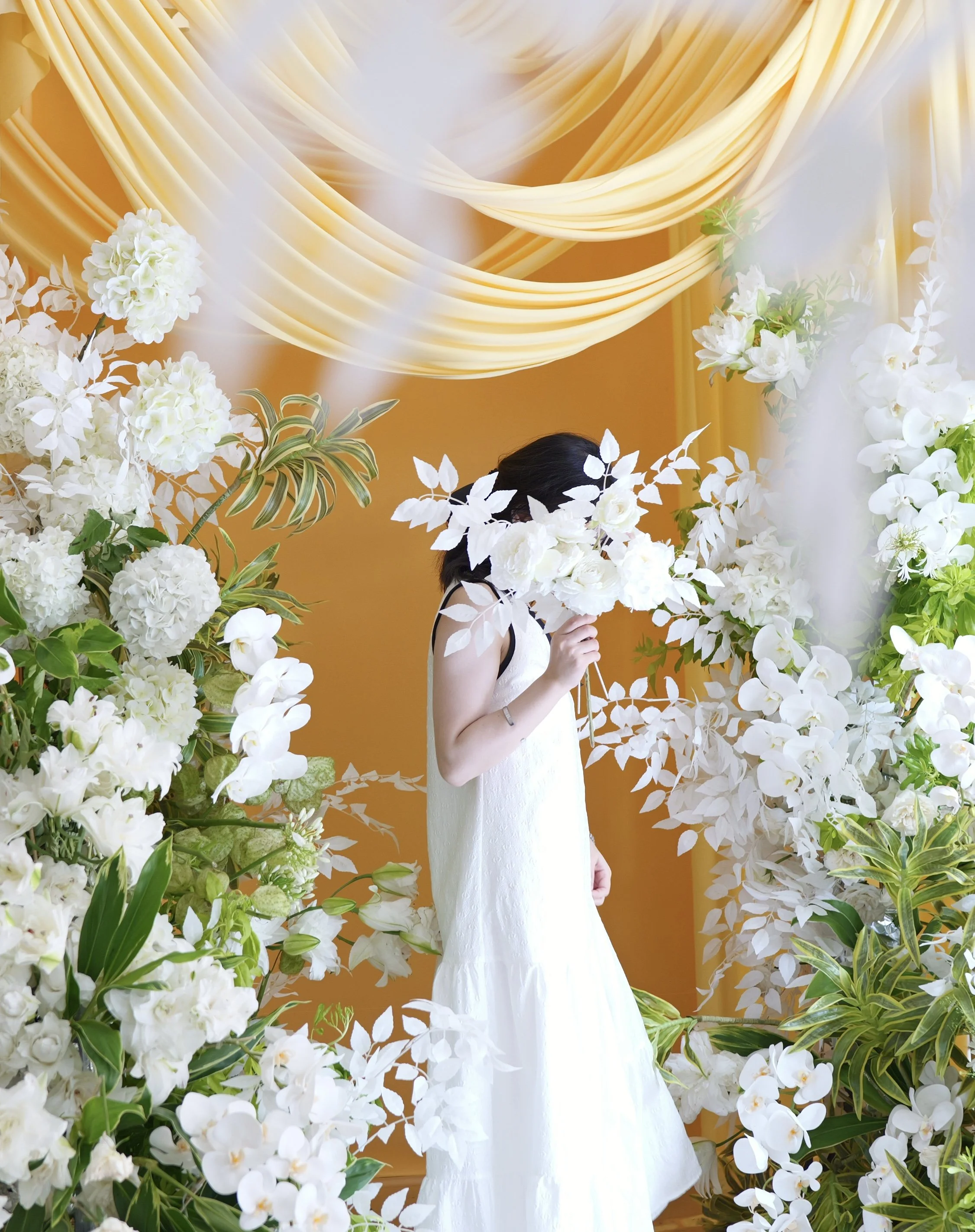A woman in a white dress standing among white flowers, holding a bouquet, with yellow drapery in the background.