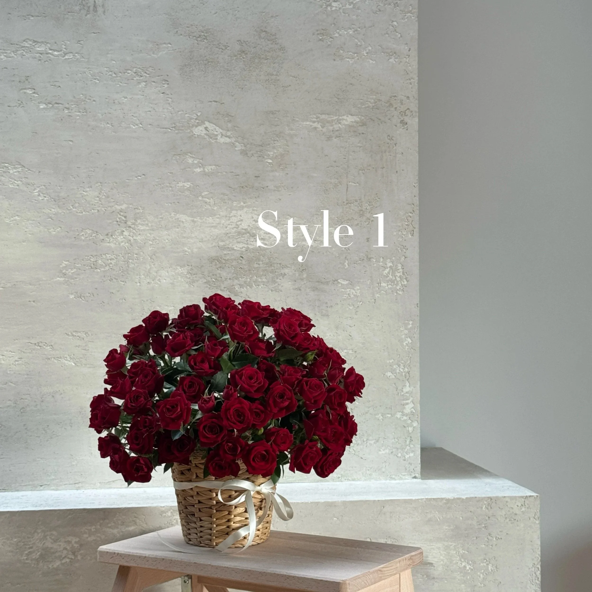 A basket of red roses with a white ribbon tied around it, placed on a wooden table with a textured off-white wall background. The text 'Style 1' is overlaid on the wall.