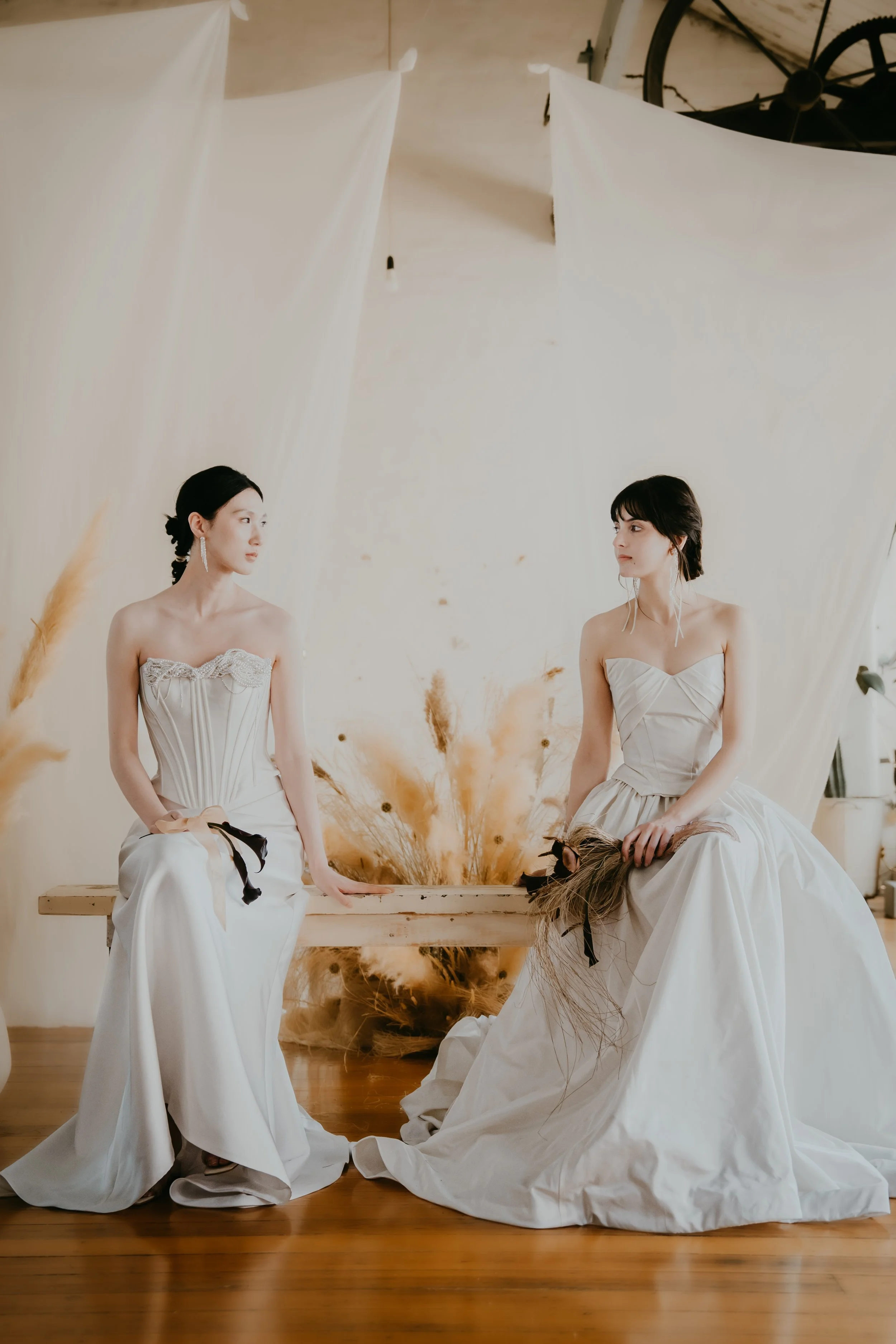 Two women in white dresses sitting on a wooden bench with a backdrop of dried grass and neutral fabric panels.