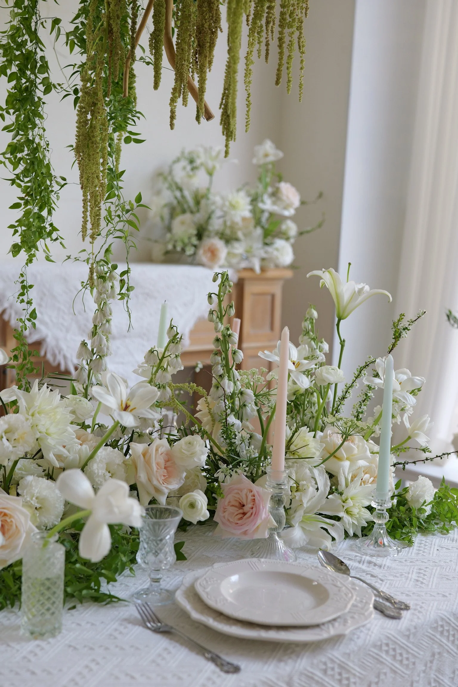 Elegant dining table with white and blush pink floral centerpiece, candles, plates, and silverware, set in a bright room with hanging greenery and floral arrangements in the background.