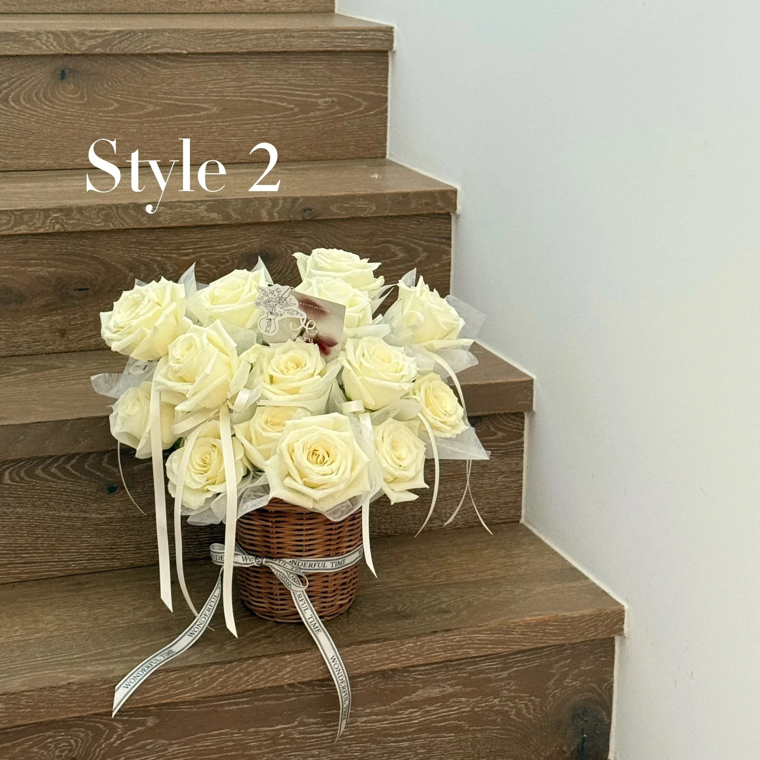 A wicker basket filled with ivory roses on a wooden staircase with a white wall on the side. The basket has a white ribbon with text and a small card attached. Text 'Style 2' is overlaid on the image.