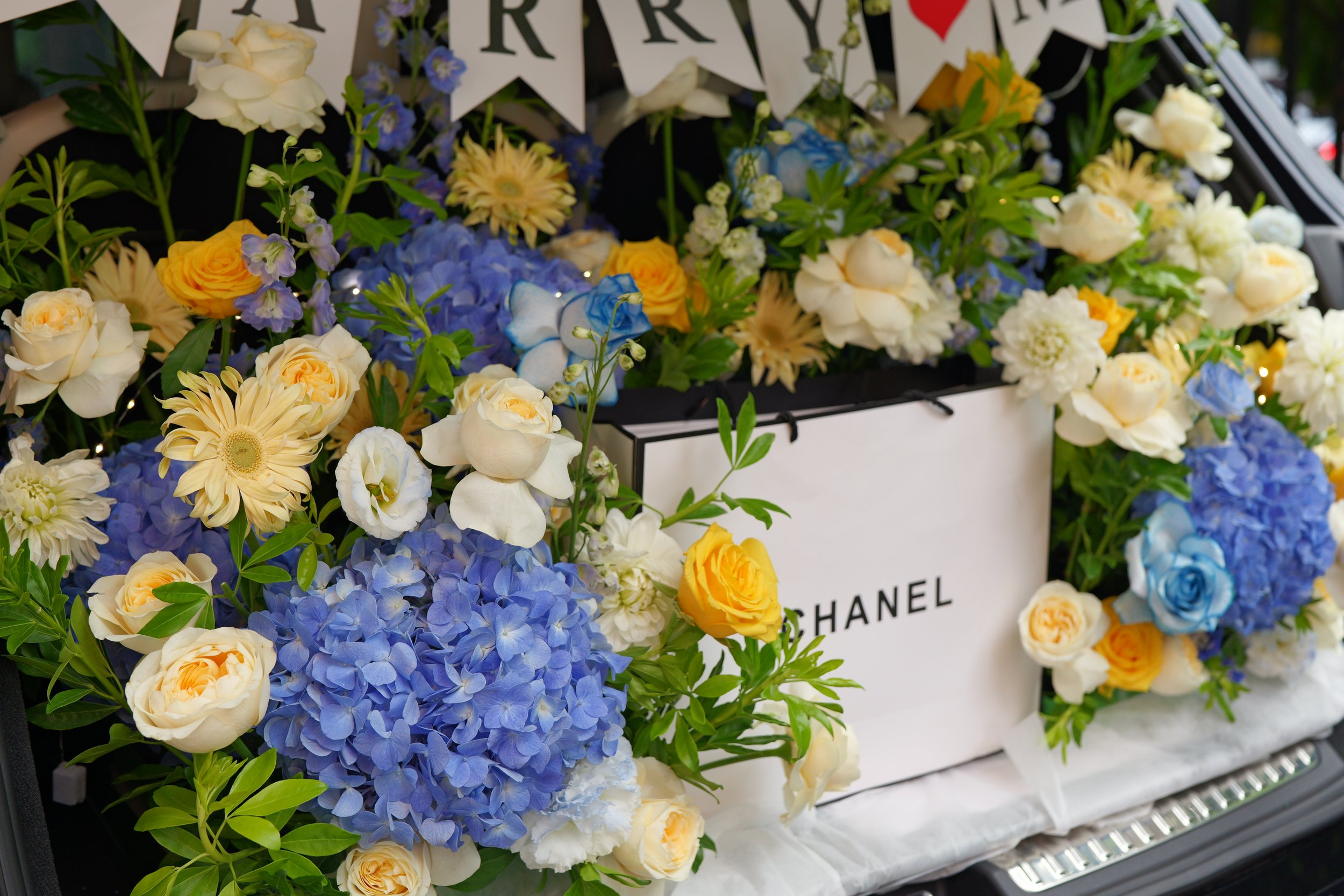 A display of mixed flowers including blue hydrangeas, white roses, cream-colored dahlias, and yellow roses, with a Chanel shopping bag among them.