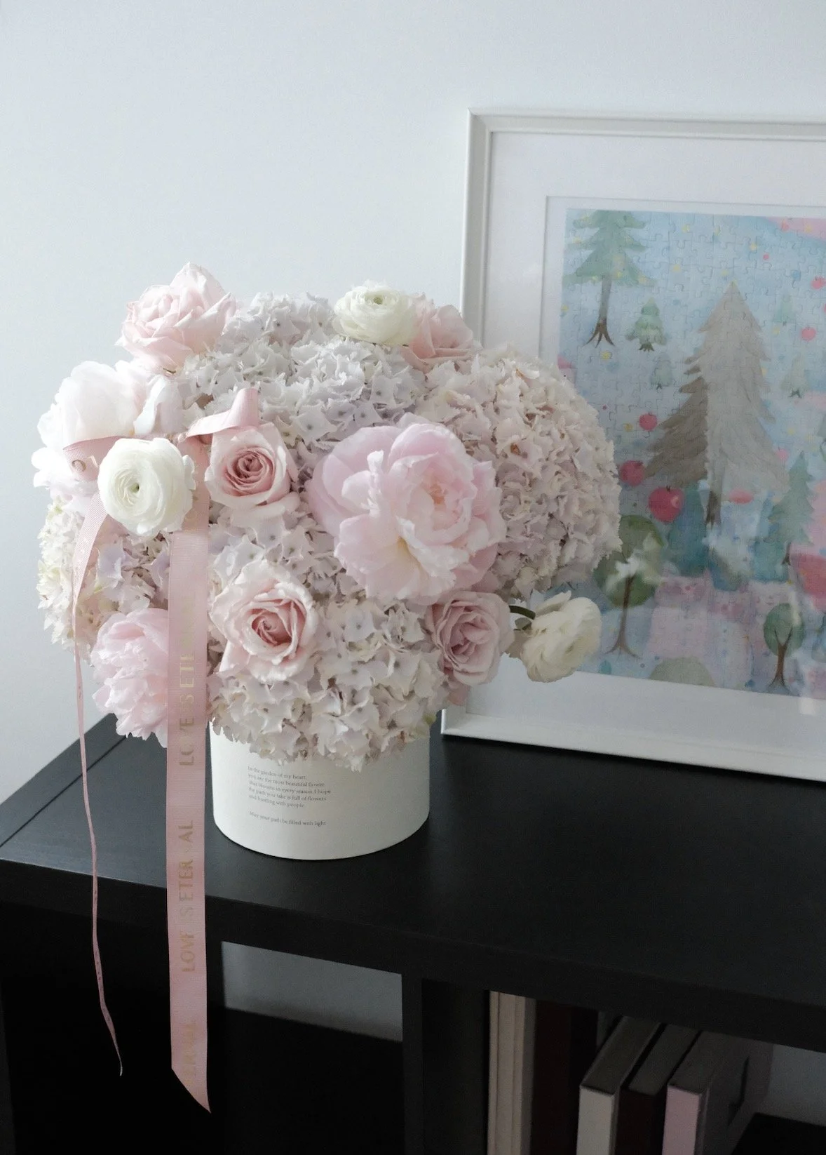 A white cylindrical vase filled with a round bouquet of pink and white flowers, including roses and hydrangeas, with a light pink ribbon attached, placed on a black table next to a framed watercolor picture of trees and a colorful background.