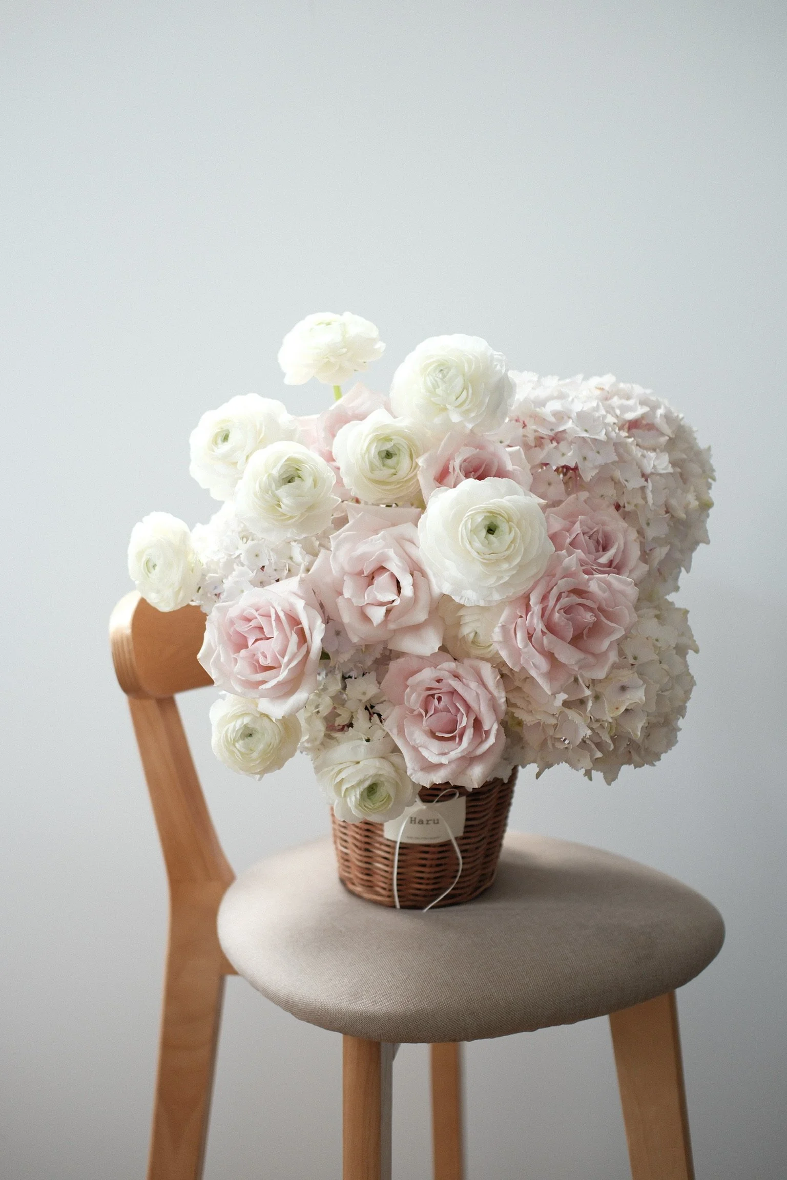 A bouquet of white and pale pink roses and white hydrangeas in a woven basket placed on a beige upholstered chair with wooden legs, against a plain white wall background.