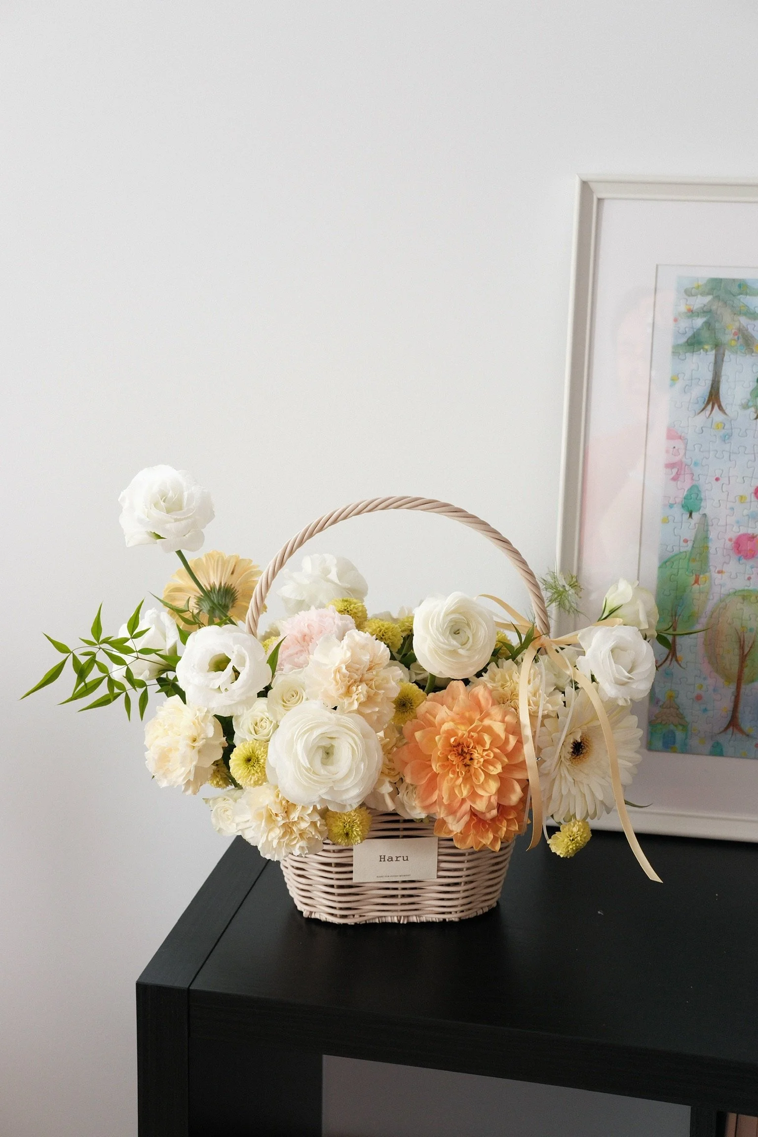 A woven basket filled with white, peach, and cream flowers, sitting on a black table.