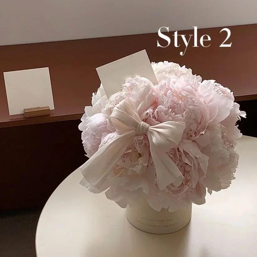 A bouquet of pink peonies with a white bow and a card, labeled as Style 2, on a white surface in a room with a brown wall.