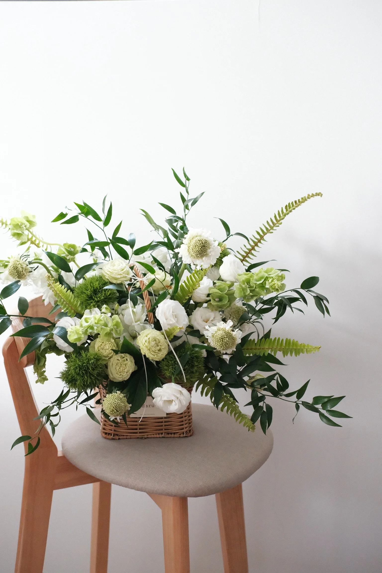 A bouquet of white and green flowers arranged in a wicker basket on a beige upholstered stool.