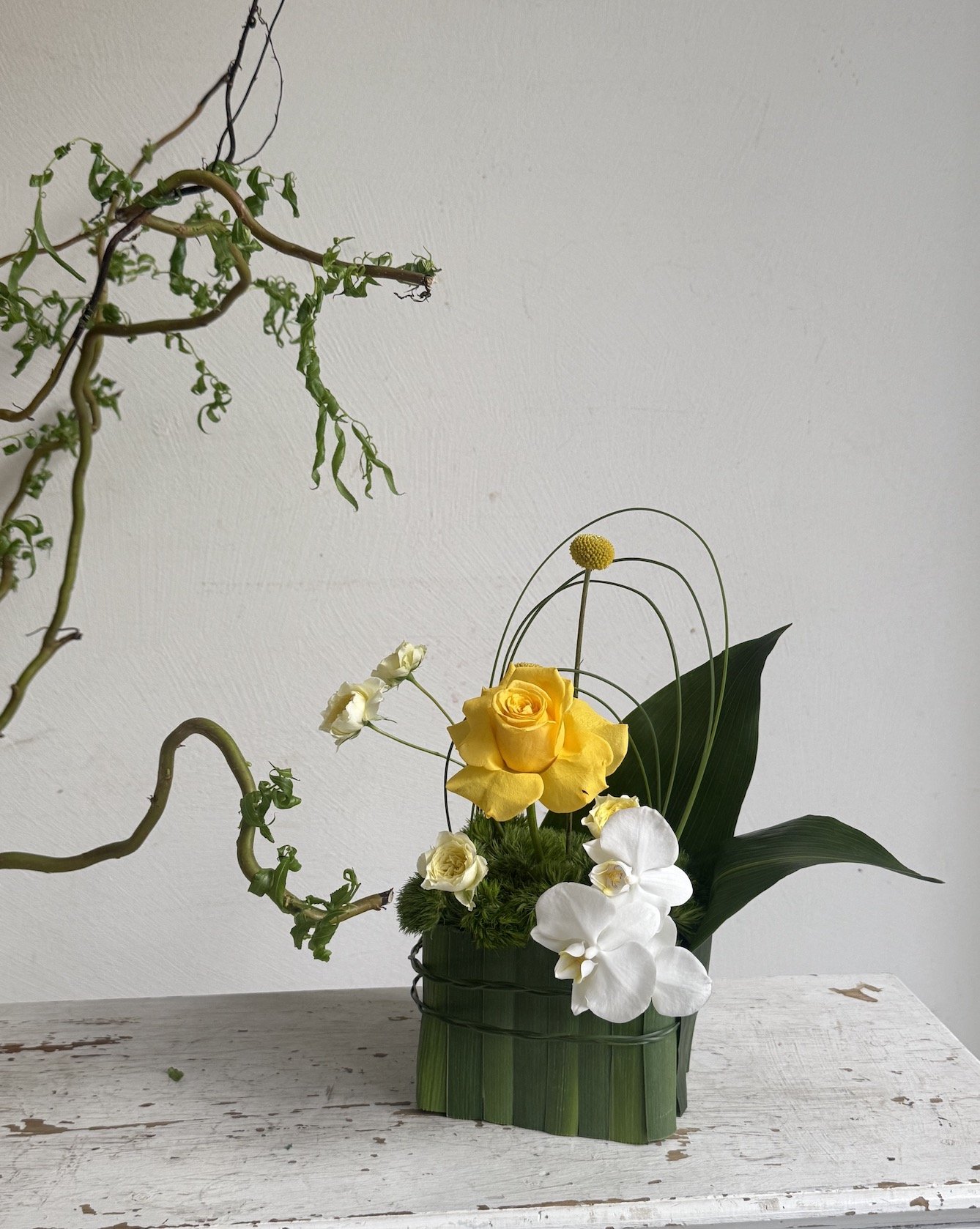 A flower arrangement with yellow rose, white orchids, and smaller white flowers in a green woven basket, with a large green leaf, on a distressed white table against a plain white wall.