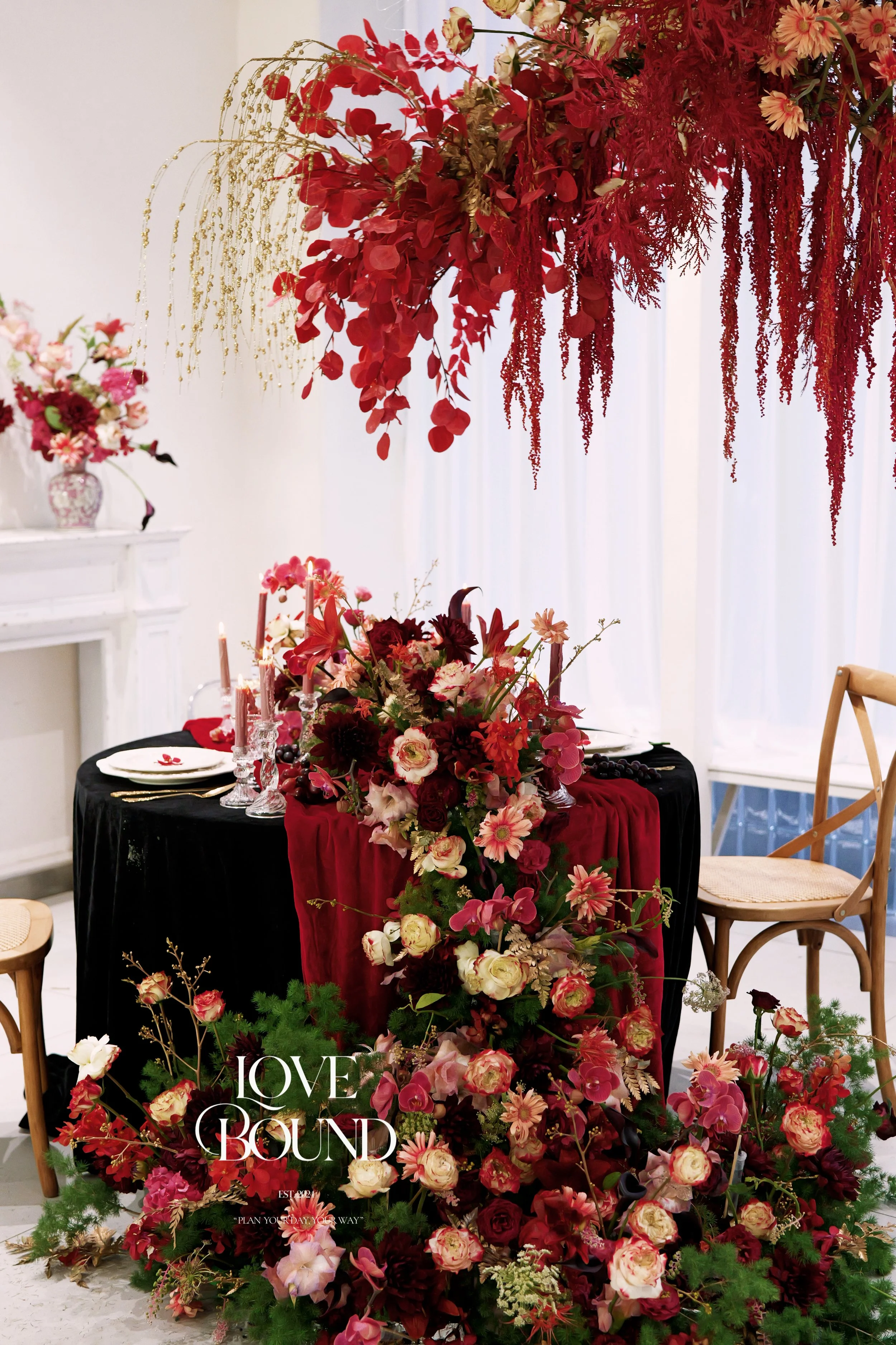 A beautifully decorated table for a romantic event with a black tablecloth and a large floral centerpiece featuring pink, red, and white flowers, surrounded by candles and chairs. Overhead, there is an elaborate hanging floral arrangement with red and pink flowers and greenery, creating an elegant and romantic atmosphere.