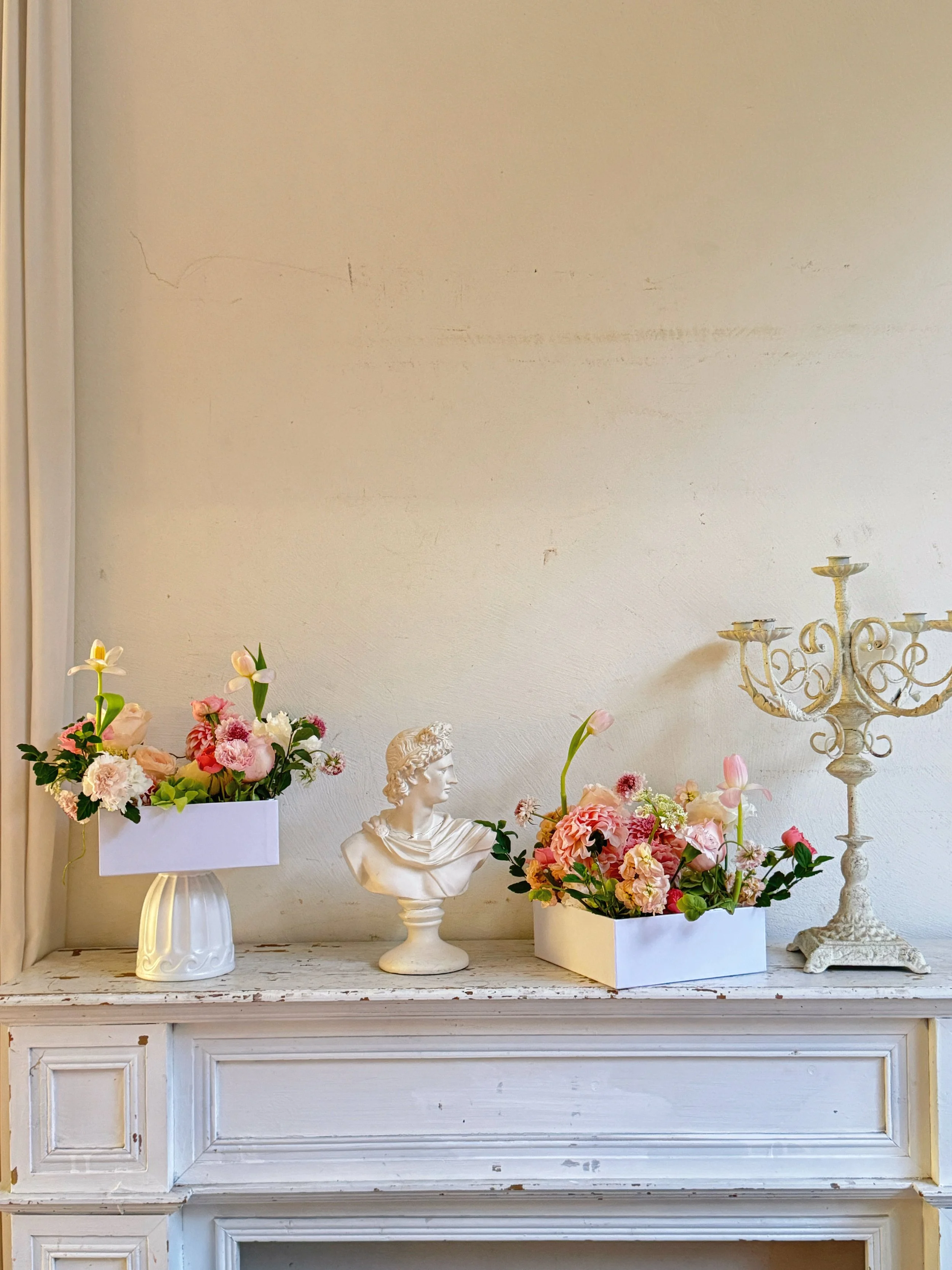 Decorative white vintage fireplace mantel with flower arrangements in white boxes, a bust sculpture, and a candle holder on a white wall.