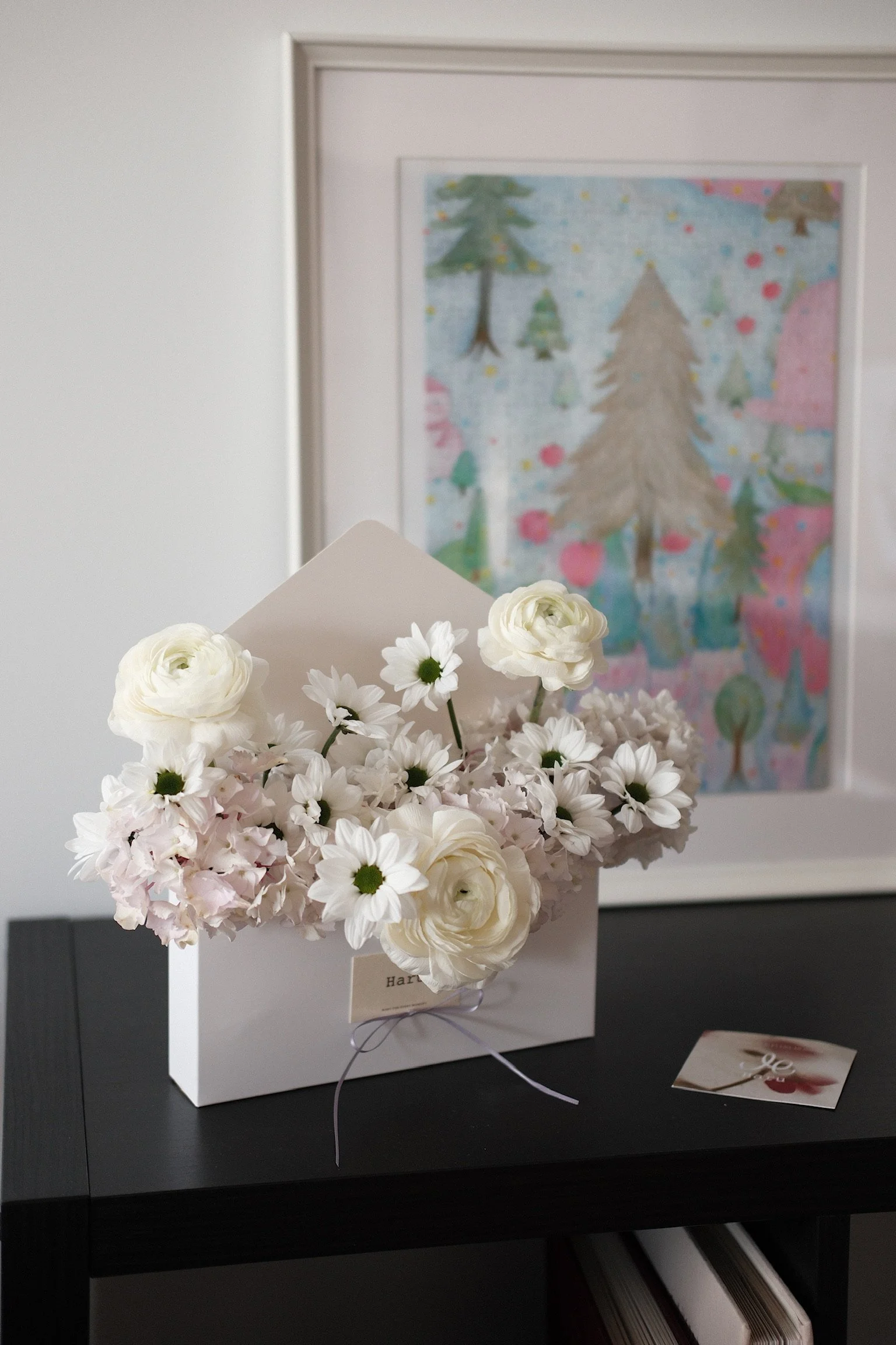 White flower bouquet in a white paper wrap on a black table with a colorful drawing of trees in the background.
