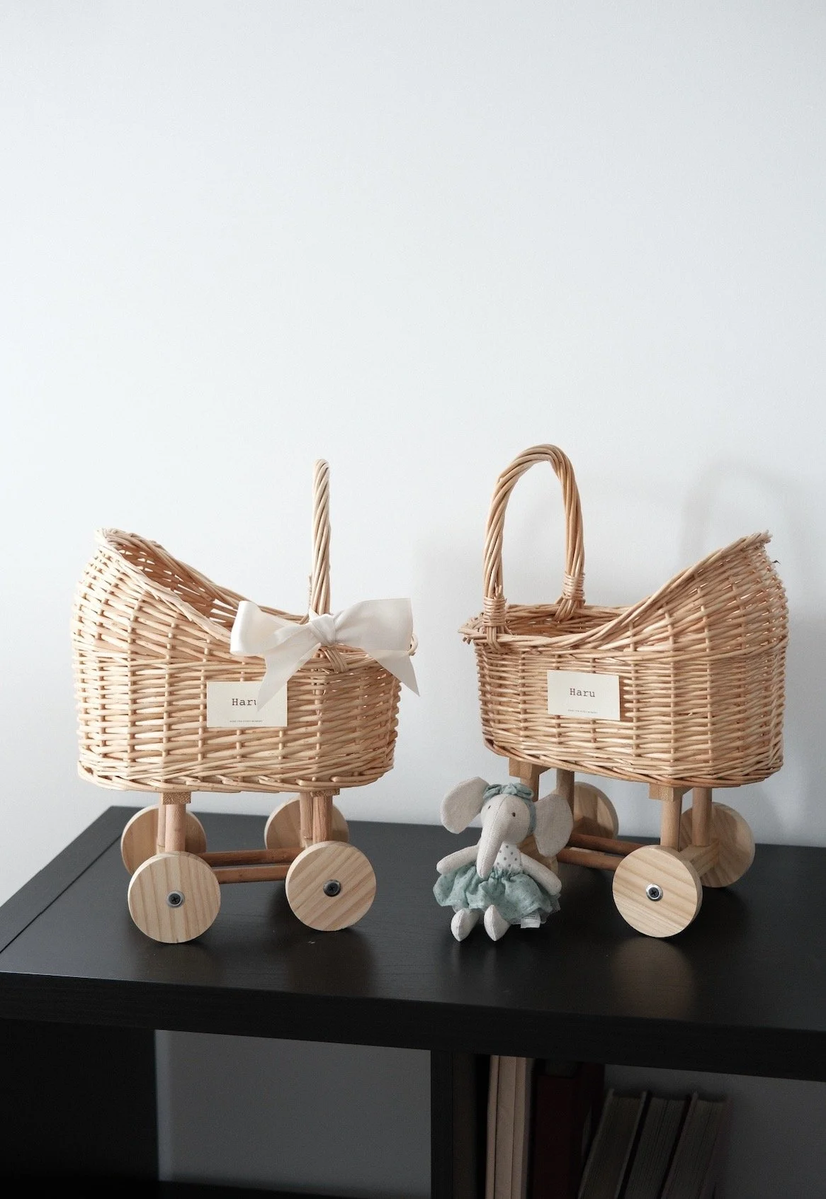 Two wicker baby strollers on a black table with a stuffed elephant toy between them, against a plain white wall. The stroller on the left has a white bow, and both have small tags with the name "Haru."
