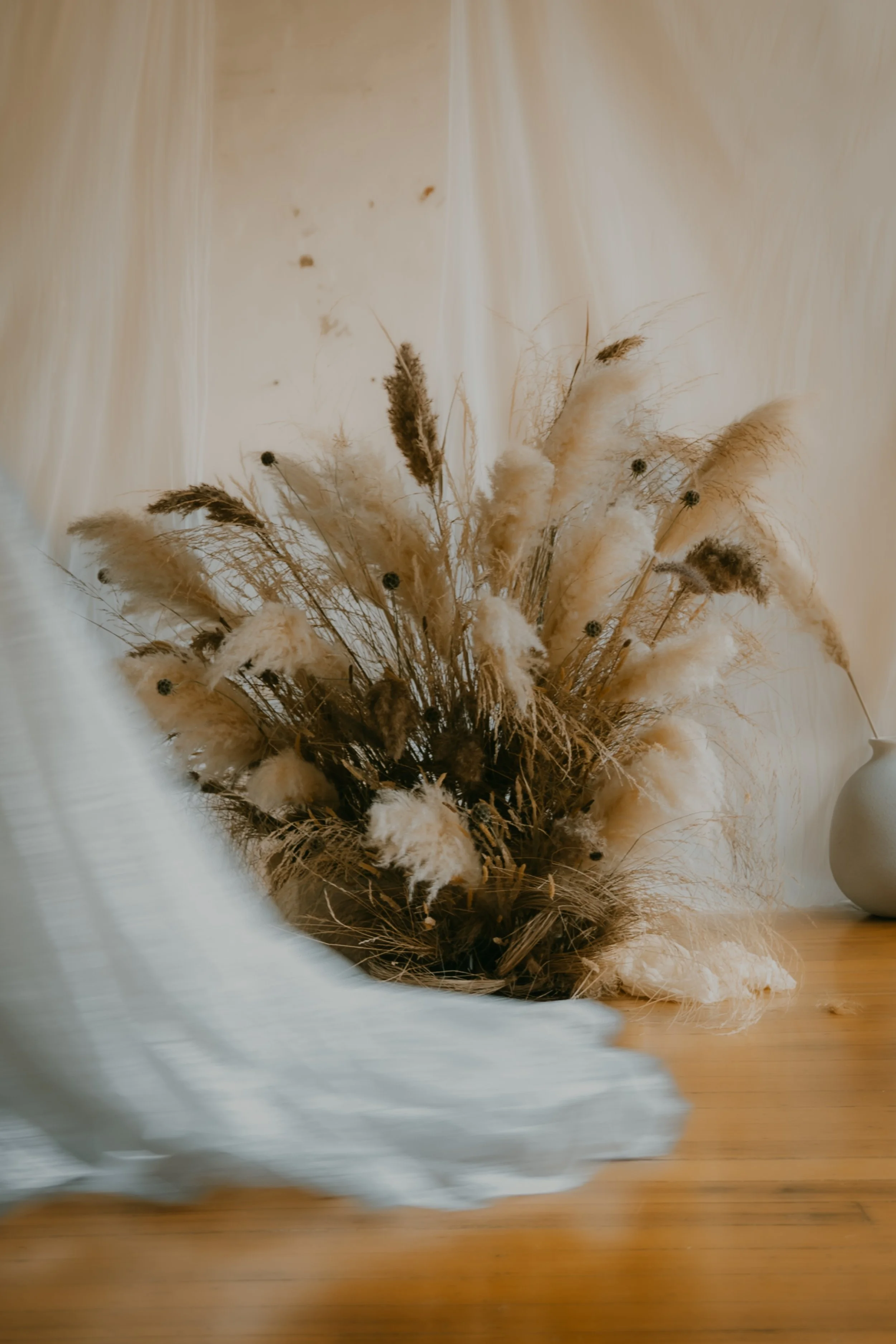 A bouquet of dried pampas grass and other dried flowers resting on a wooden surface next to a round, white vase.
