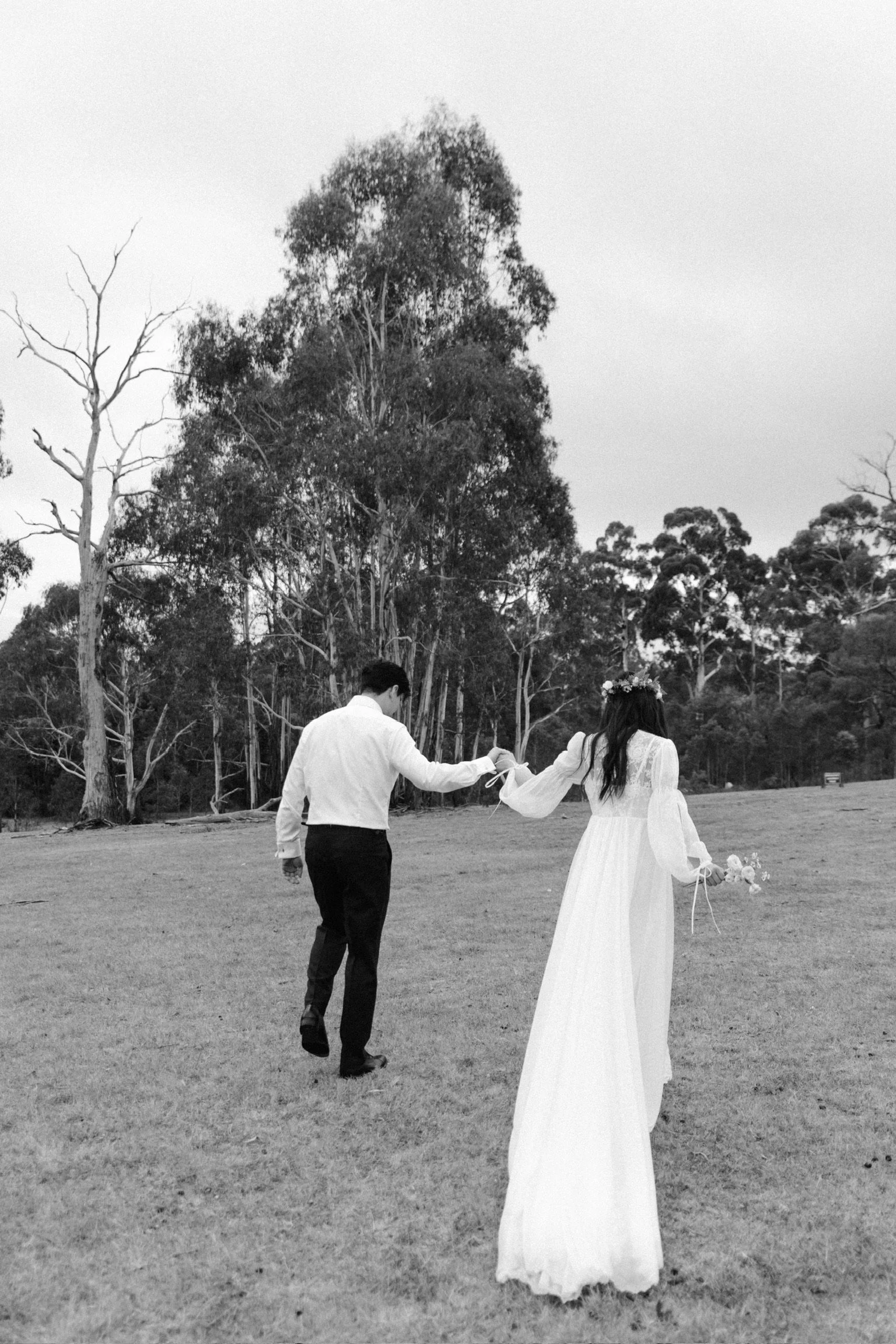 A black and white photo of a couple holding hands and walking away in a field, with trees in the background. The woman is wearing a long, flowing dress and flower crown, holding a bouquet, while the man is dressed in a white shirt and dark pants.