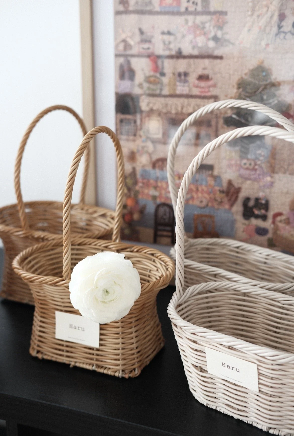 Three woven baskets with handles, two in a natural wood color and one painted white, sit on a black surface with a white flower attached to one basket. Each basket has a label with the word 'Haru.' A blurred colorful background hangs on the wall behind them.