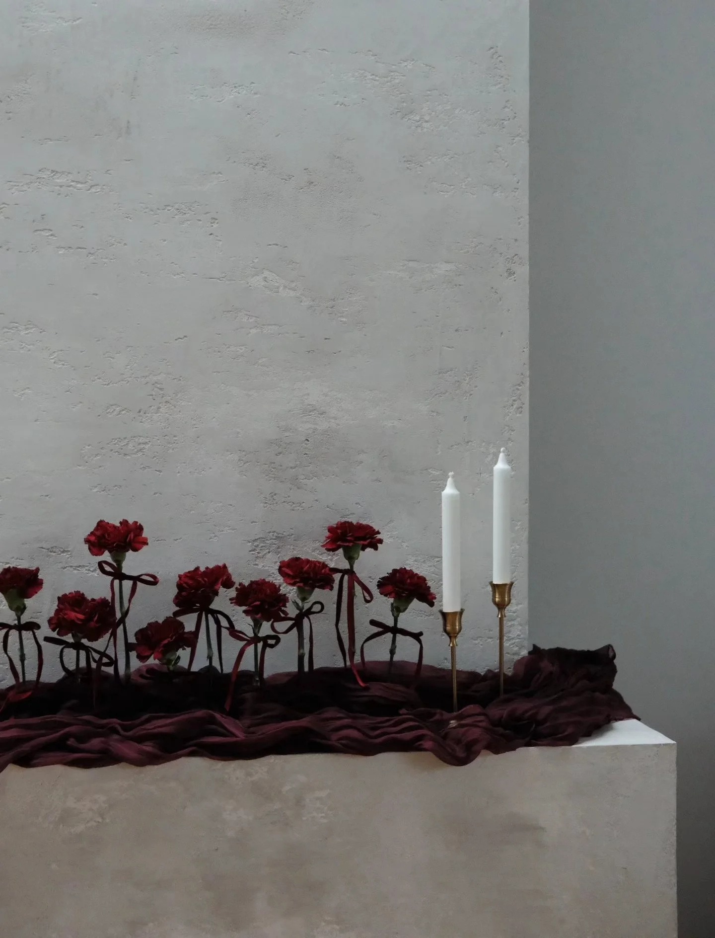 Decorative display with red flowers tied with ribbons and two white candles in holders on dark fabric.