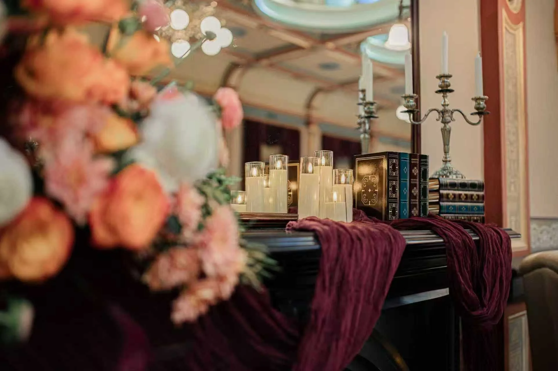 A view of a decorated room with a table draped in velvet fabric, several lit candles, a floral arrangement on the left, and books near a wall sconce in the background.