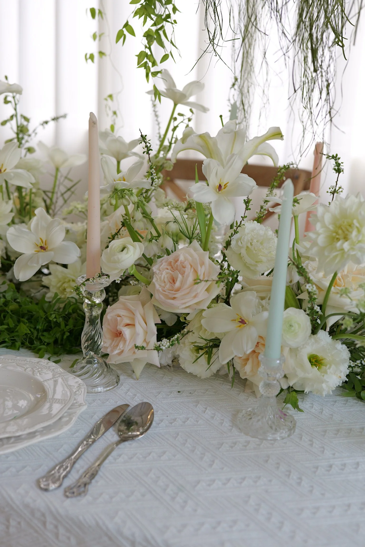 Elegant table setting with a floral centerpiece consisting of white and blush roses, lilies, and greenery, accented by pastel-colored candles in glass holders, on a white textured tablecloth, with a blurred background of a chair and curtains.