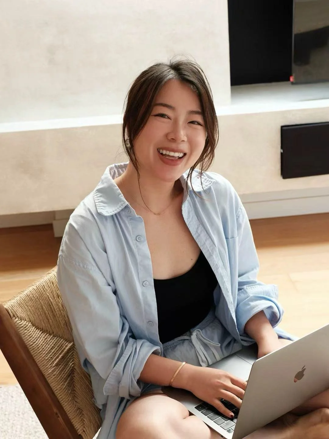 A woman smiling while sitting on a chair with a laptop on her lap, wearing a light blue shirt and black top, in a room with beige walls and wooden flooring.