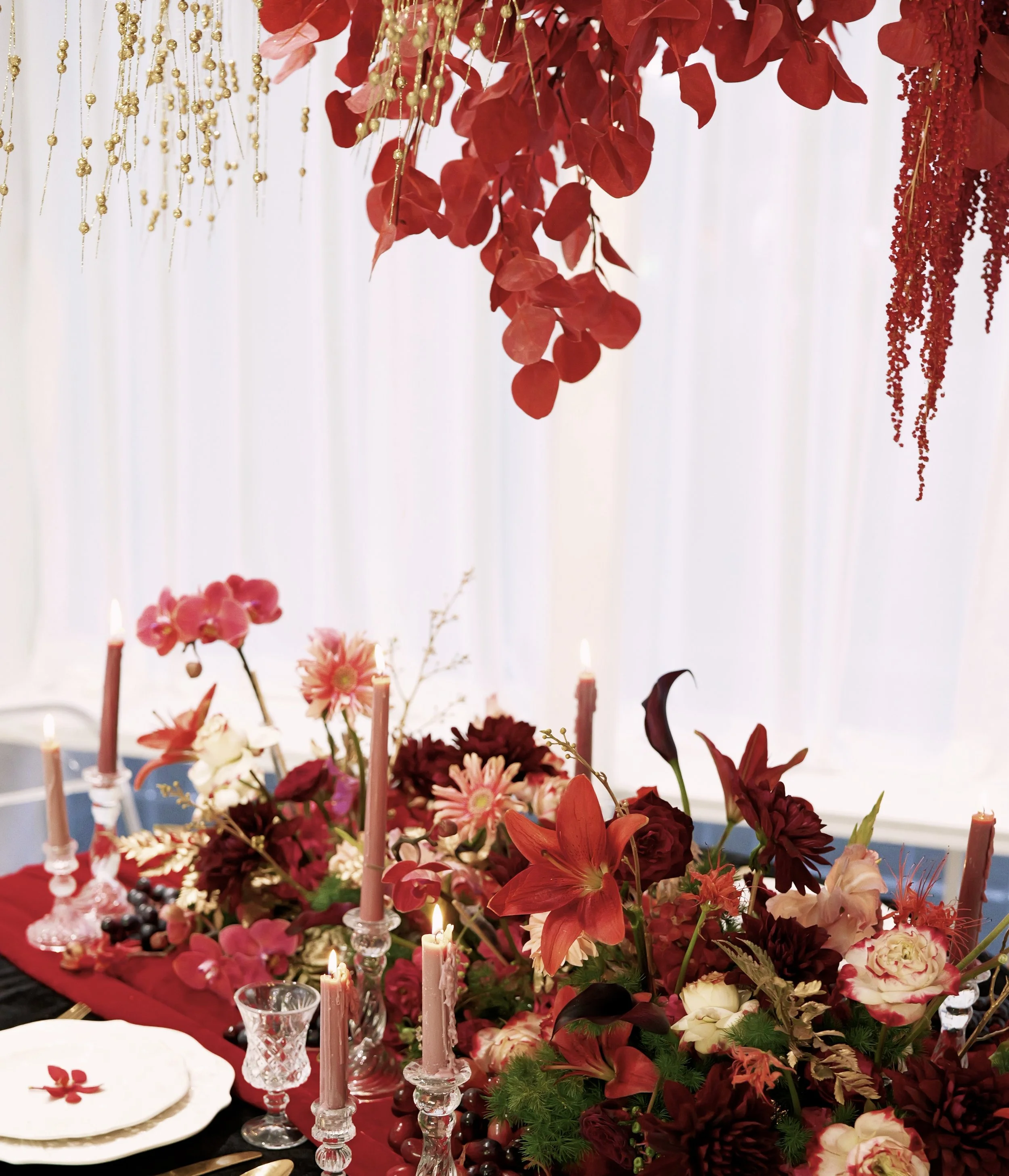 A table decorated with a lush arrangement of red, pink, and white flowers, tall pink candles in glass holders, and a red cloth underlining the display. Hanging above are red leaves and gold beads against a white backdrop.