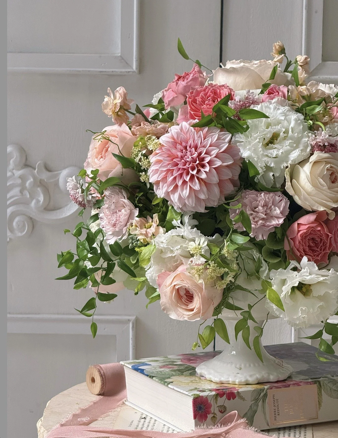 A bouquet of pink, white, and peach flowers in a white vase on top of a book, placed on a pink cloth on a table. The background features a white wall with decorative molding.