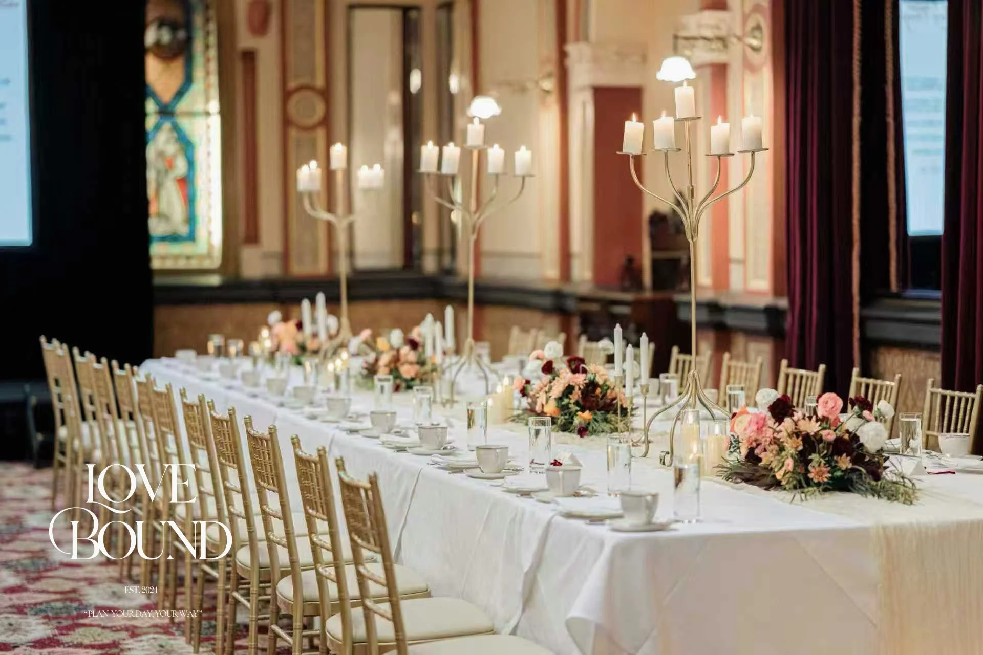 Elegant banquet table set with floral centerpieces, candles, and gold chairs in a decorated event hall.