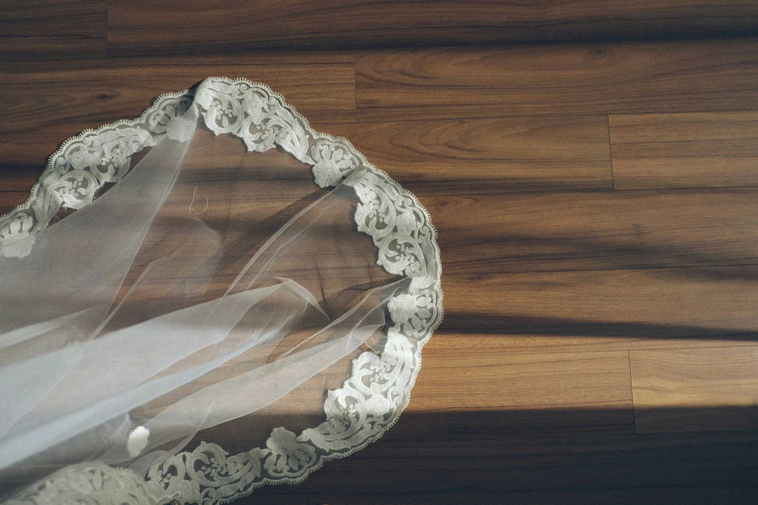 A sheer lace veil with intricate embroidery along the edge, laid on a wooden floor.