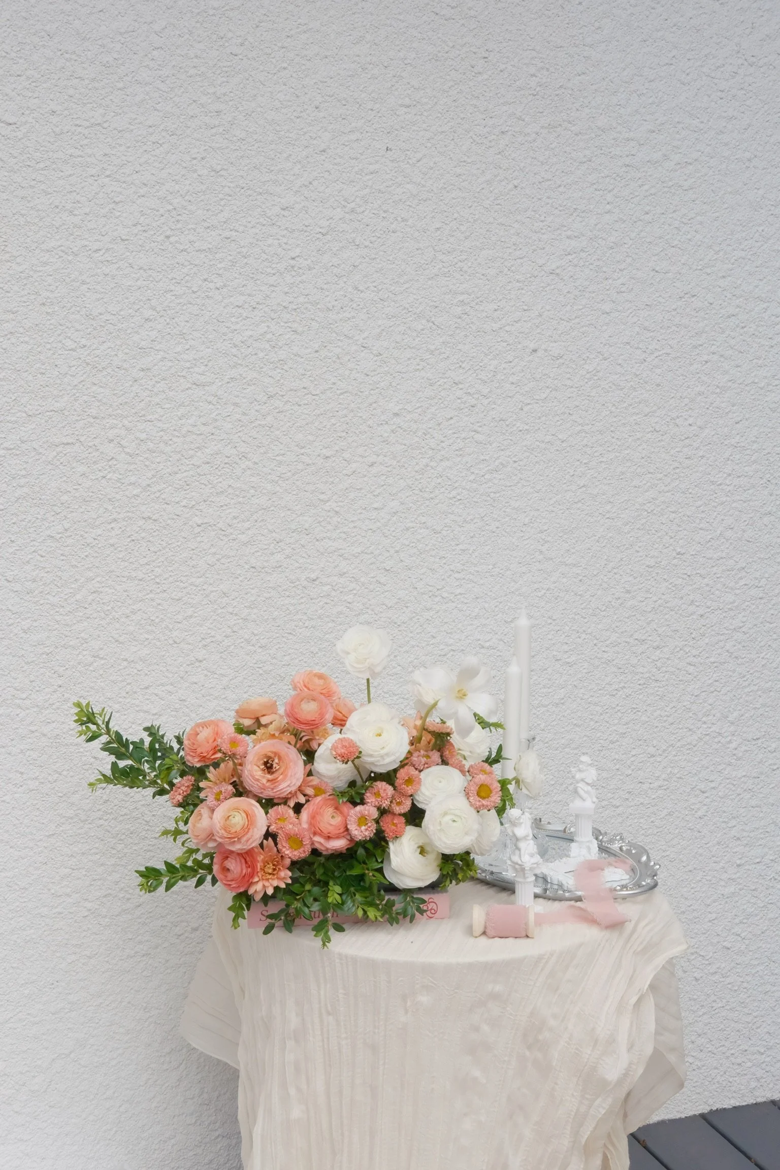 A flower arrangement with pink and white flowers and green foliage on a table covered with a white tablecloth, next to white candles and silver decorative items against a white textured wall.