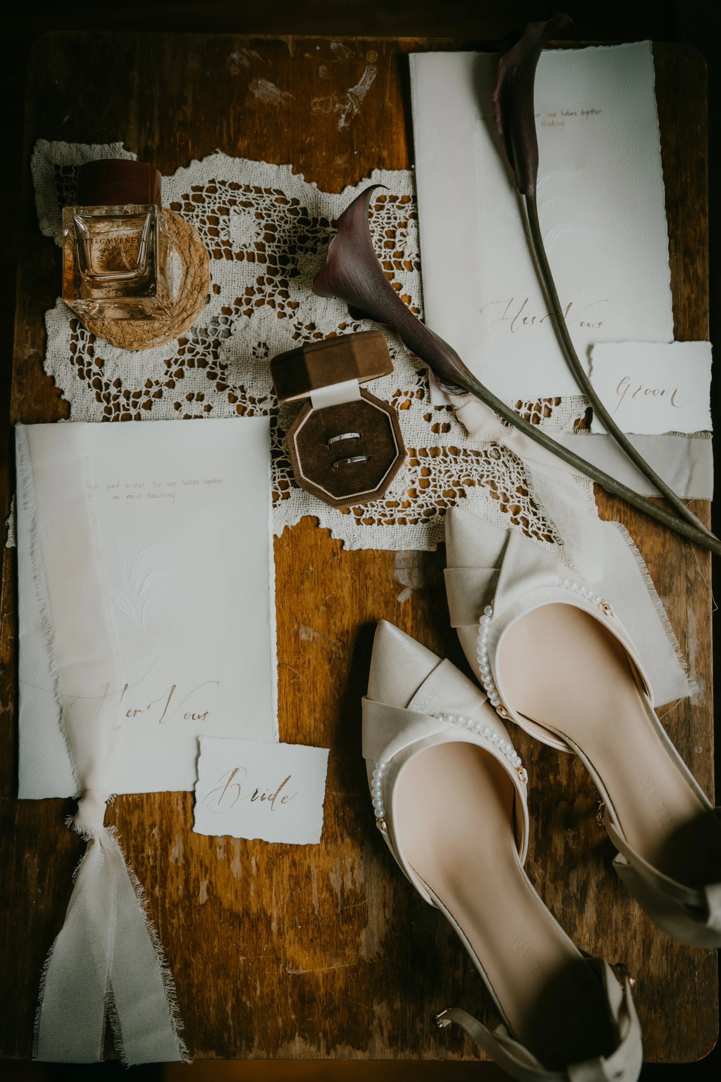 Wedding accessories on a wooden table, including white high heels with pearl details, a perfume bottle, a small box with rings, handwritten notes, calla lilies, and lace fabric.
