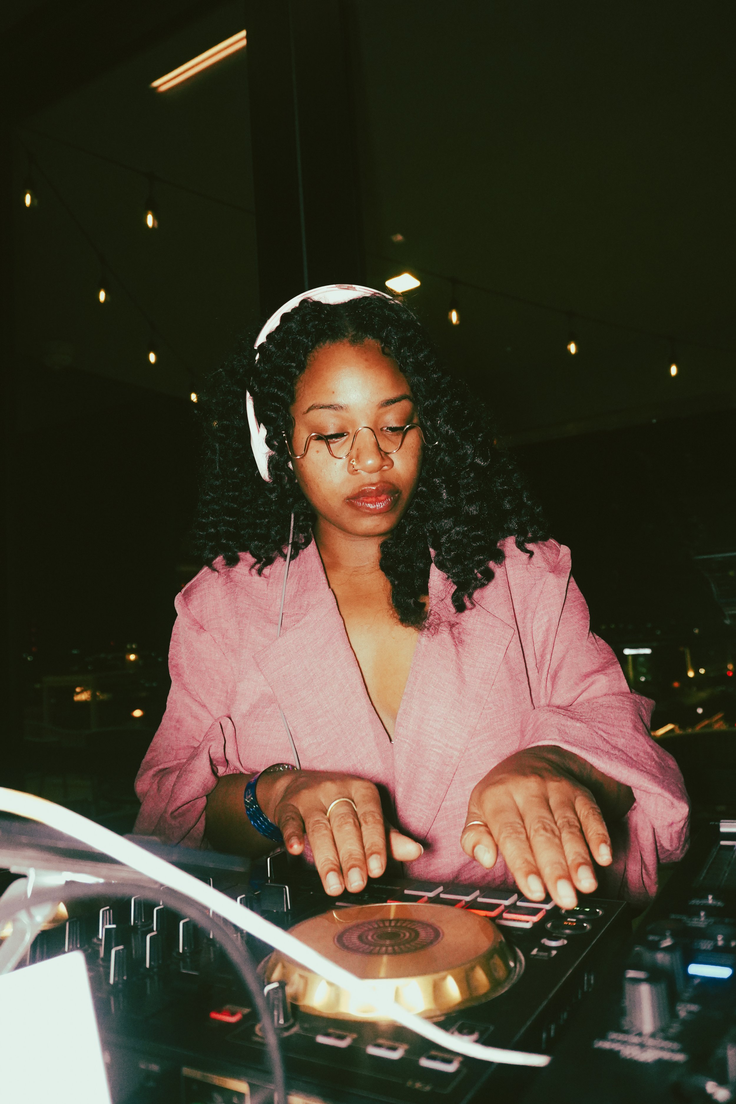 A young woman with curly black hair, glasses, and a nose ring, wearing a pink blazer, is DJing with headphones on. She is operating DJ equipment in a dimly lit environment with string lights in the background.