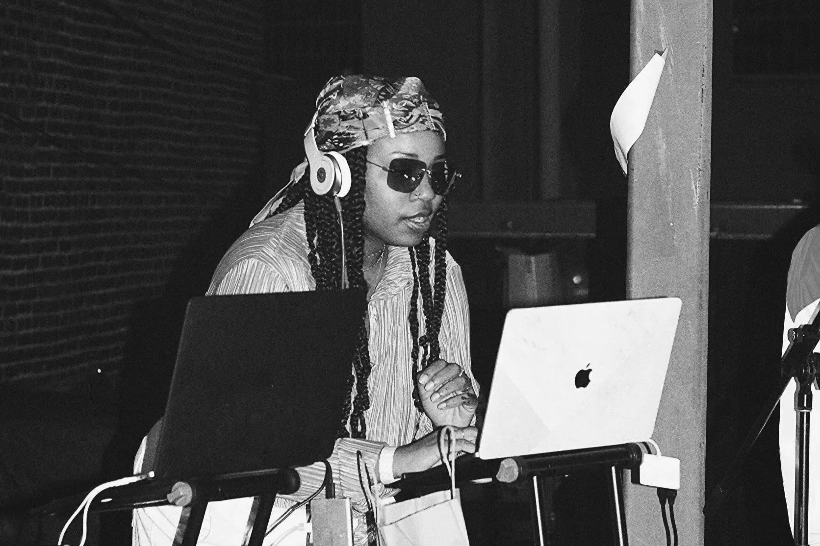 A woman with braided hair, wearing sunglasses, headphones, a patterned headscarf, and a striped shirt, working at a DJ setup with two laptops, including an Apple MacBook.