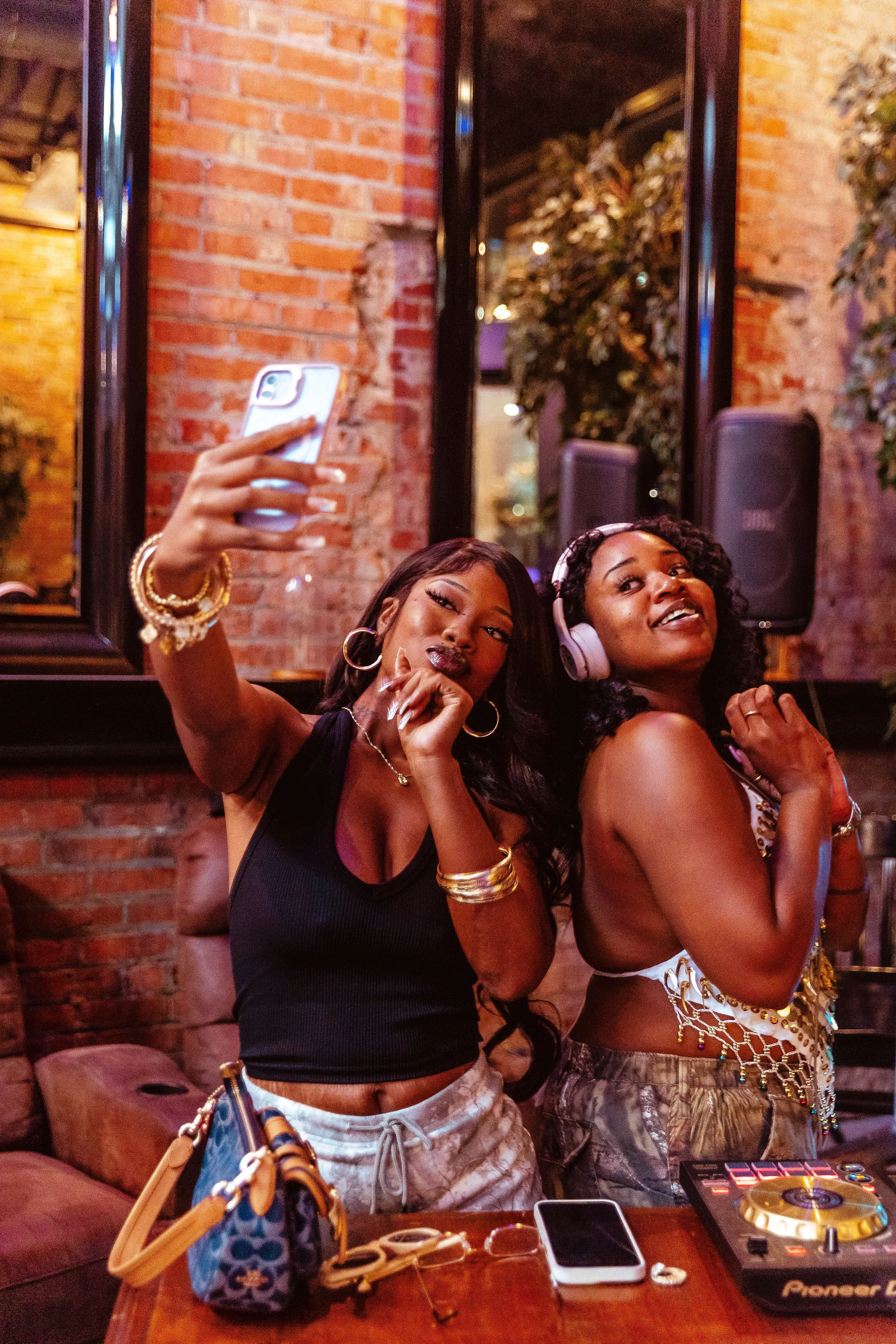 Two women at a party or club, taking a selfie, one wearing headphones and the other making a peace sign, with DJ equipment on the table.