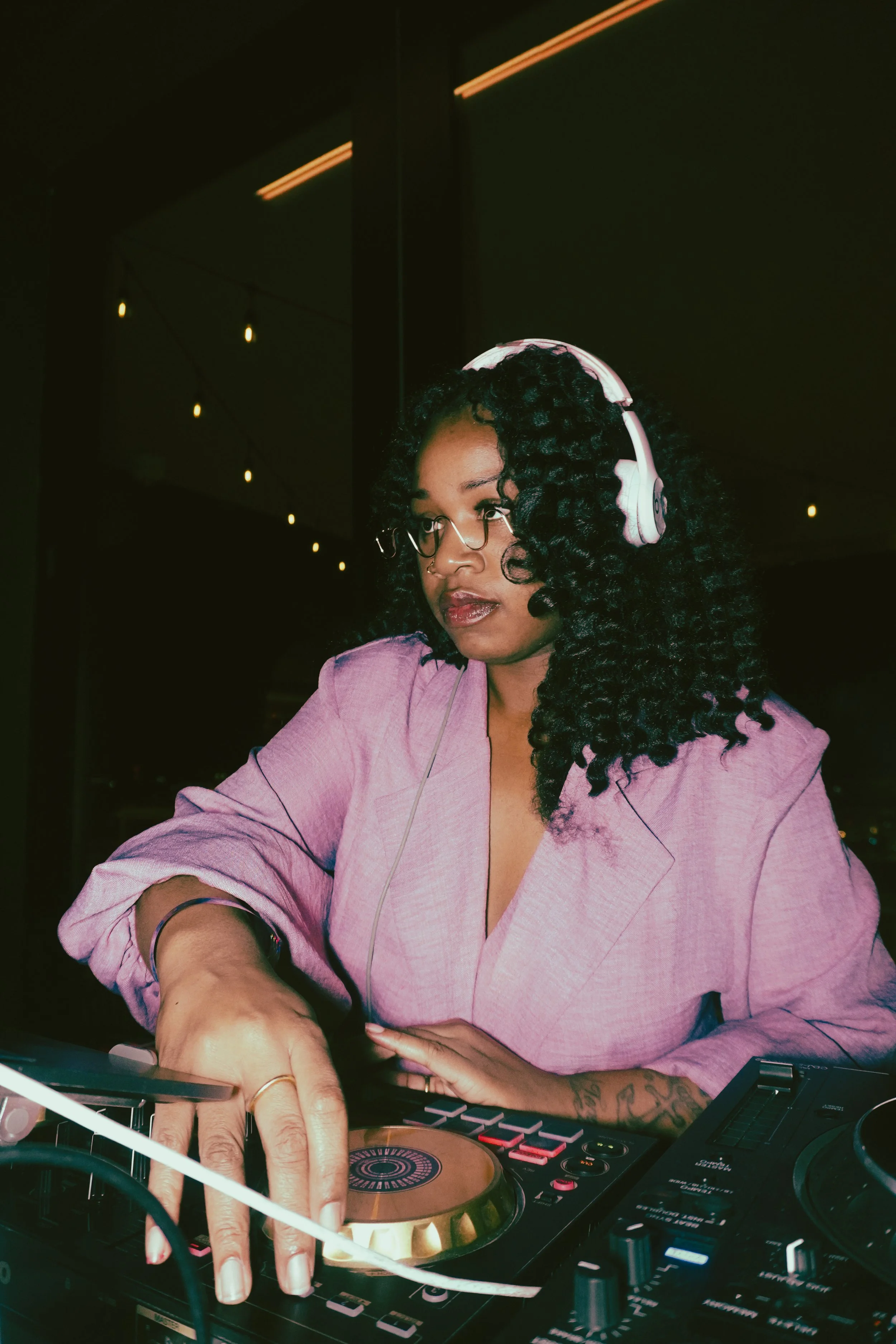A woman with curly black hair, wearing glasses, white headphones, and a pink blazer, is DJing using DJ equipment at a dimly lit venue.