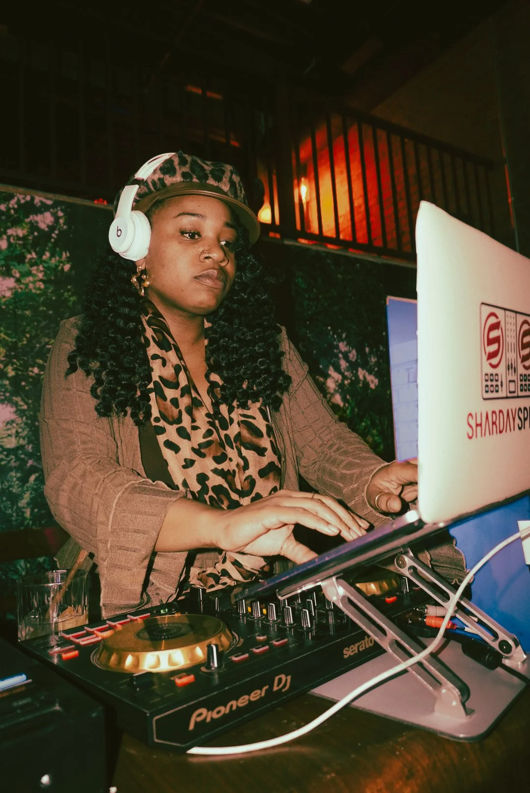 A woman DJ wearing a leopard print cap, matching scarf, and headphones, standing behind a Pioneer DJ controller and laptop setup, in a dimly lit indoor venue.