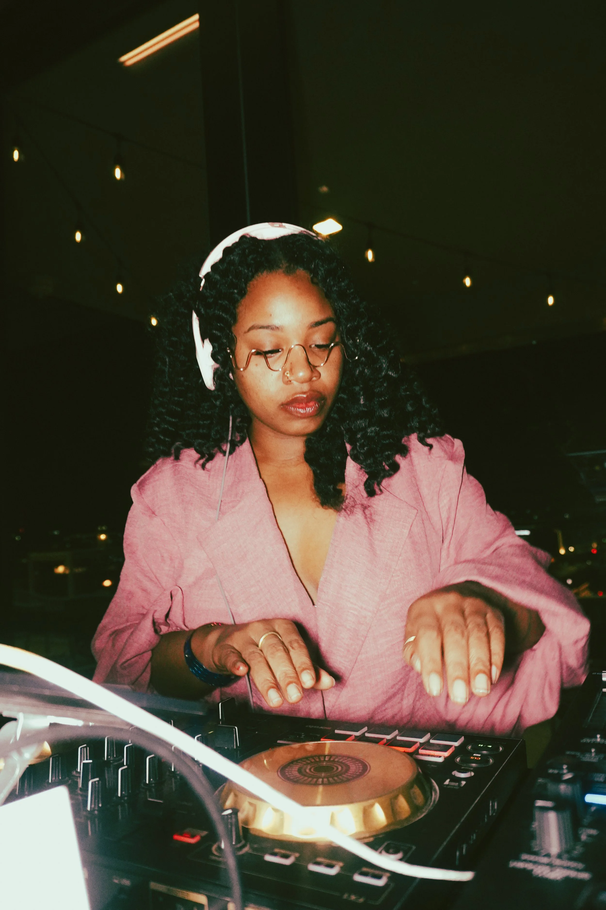 A woman with curly black hair, glasses, and a nose piercing wearing a pink blazer, DJing with headphones and a professional DJ controller in a dimly lit environment with hanging string lights.