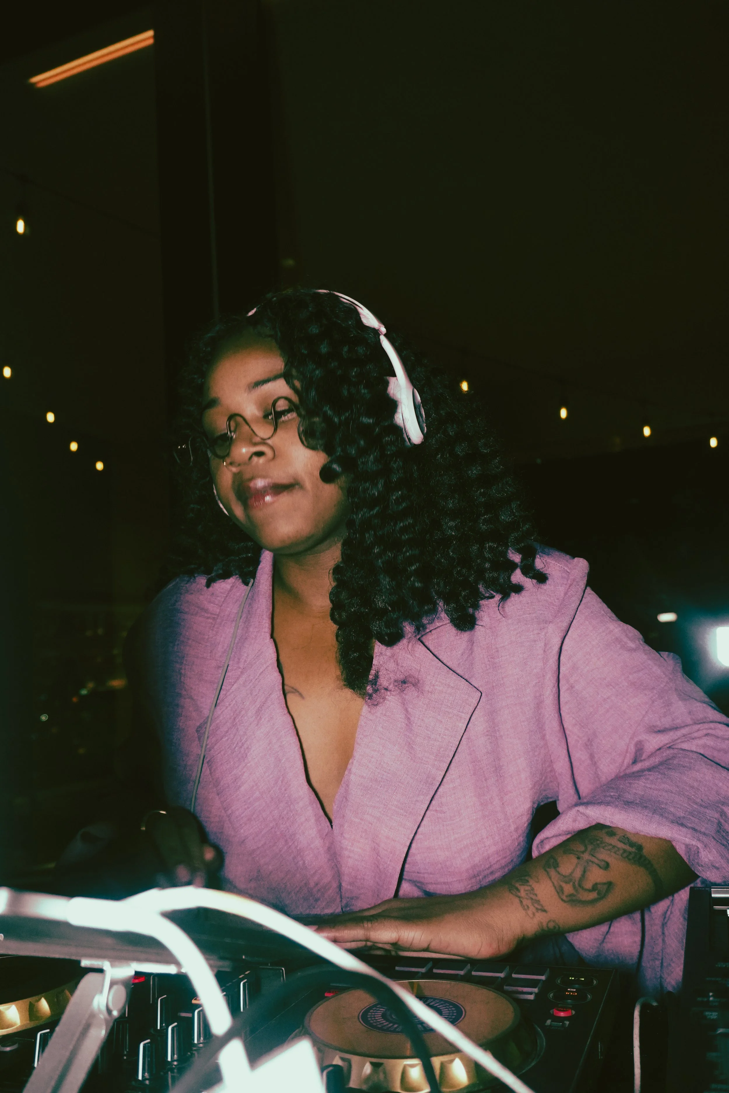 Woman DJ with curly hair, headphones, glasses, and tattoos on her arm, operating DJ equipment in a dimly lit room.