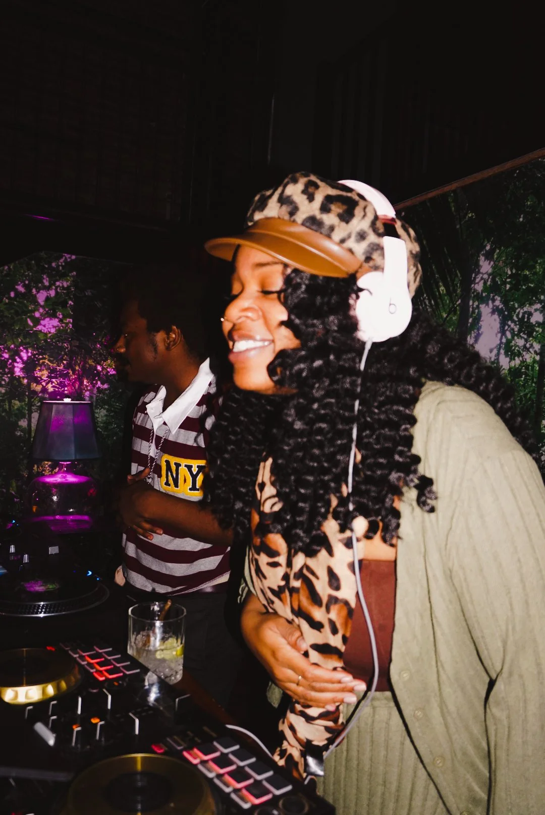 Two women at a DJ booth enjoying music. The woman in front is wearing a leopard print hat, headphones, and a beige jacket with cheetah print, smiling with eyes closed. The woman behind has dark curly hair and is wearing a white and maroon striped shi