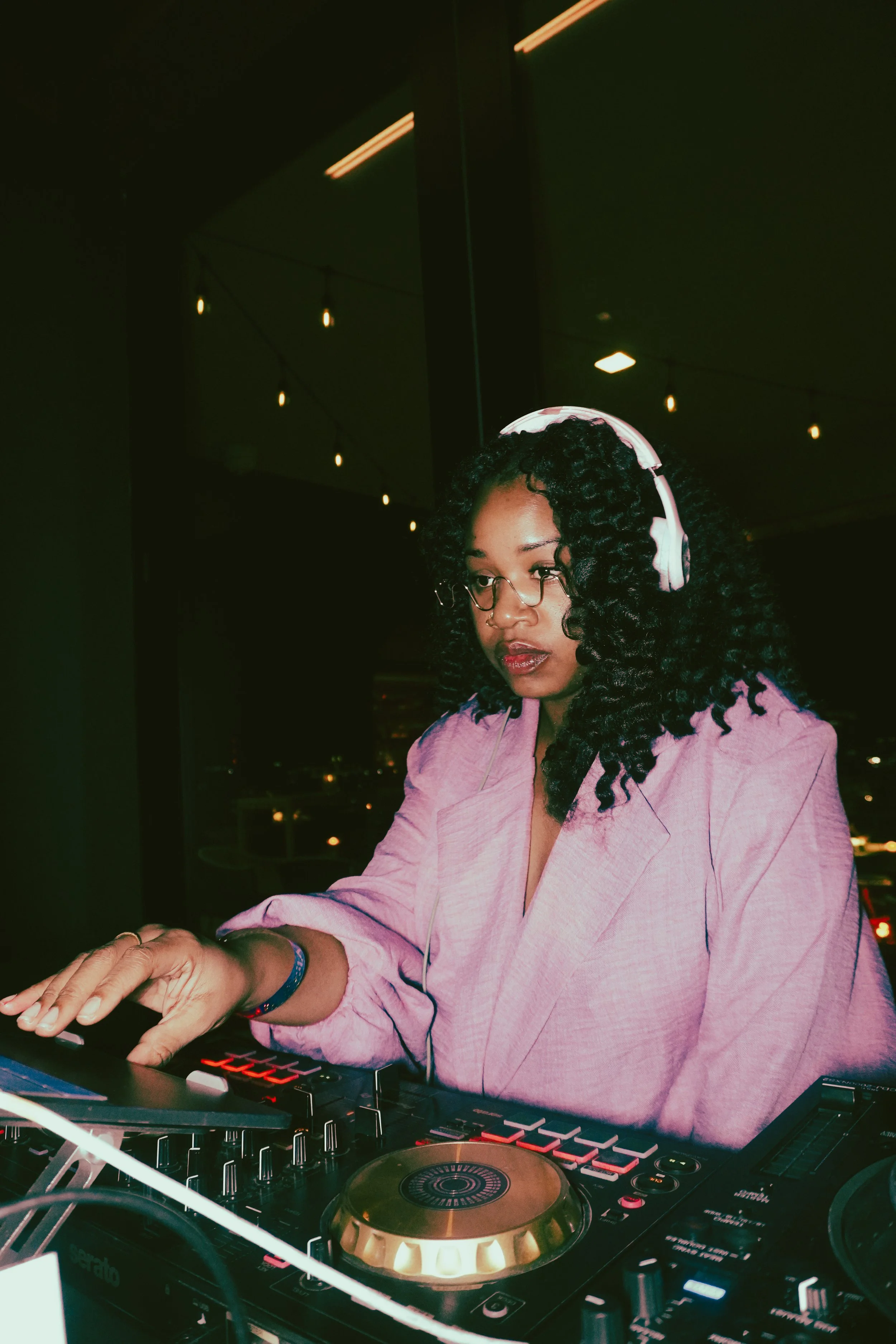 A woman with curly black hair, glasses, and a pink blazer, DJing with headphones on in a nighttime setting.